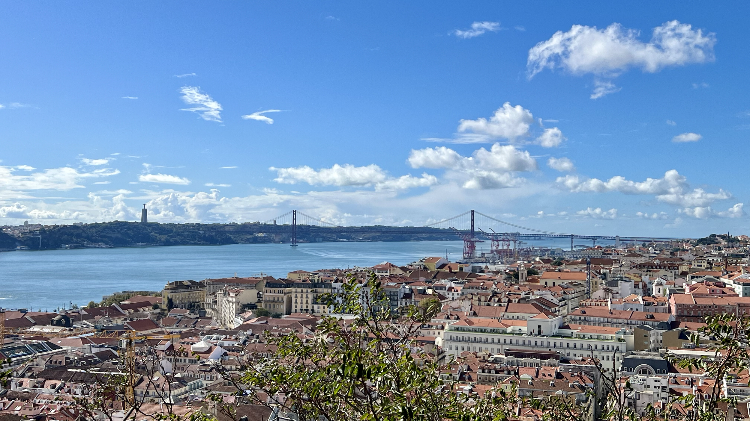 A cityscape with red-roofed buildings, a river with a suspension bridge, and a statue on a hill in the background under a partly cloudy sky.