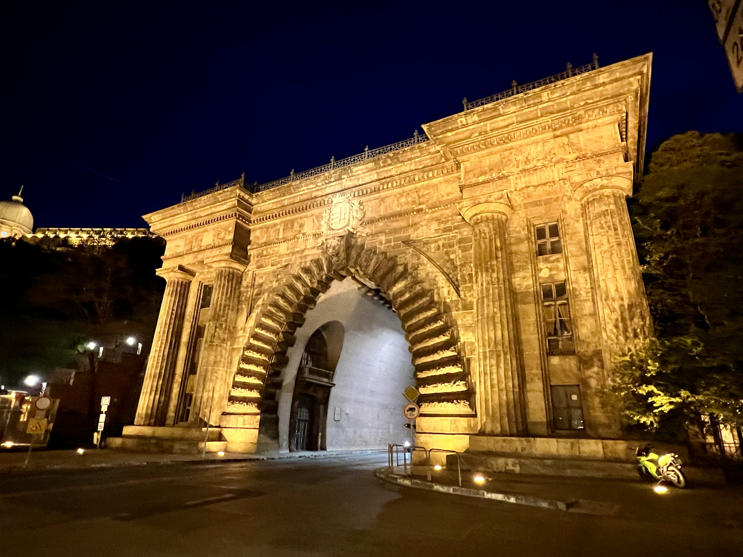 Night view of an illuminated historic stone arch gateway with classical columns and detailed architecture, with trees and a dark sky in the background.