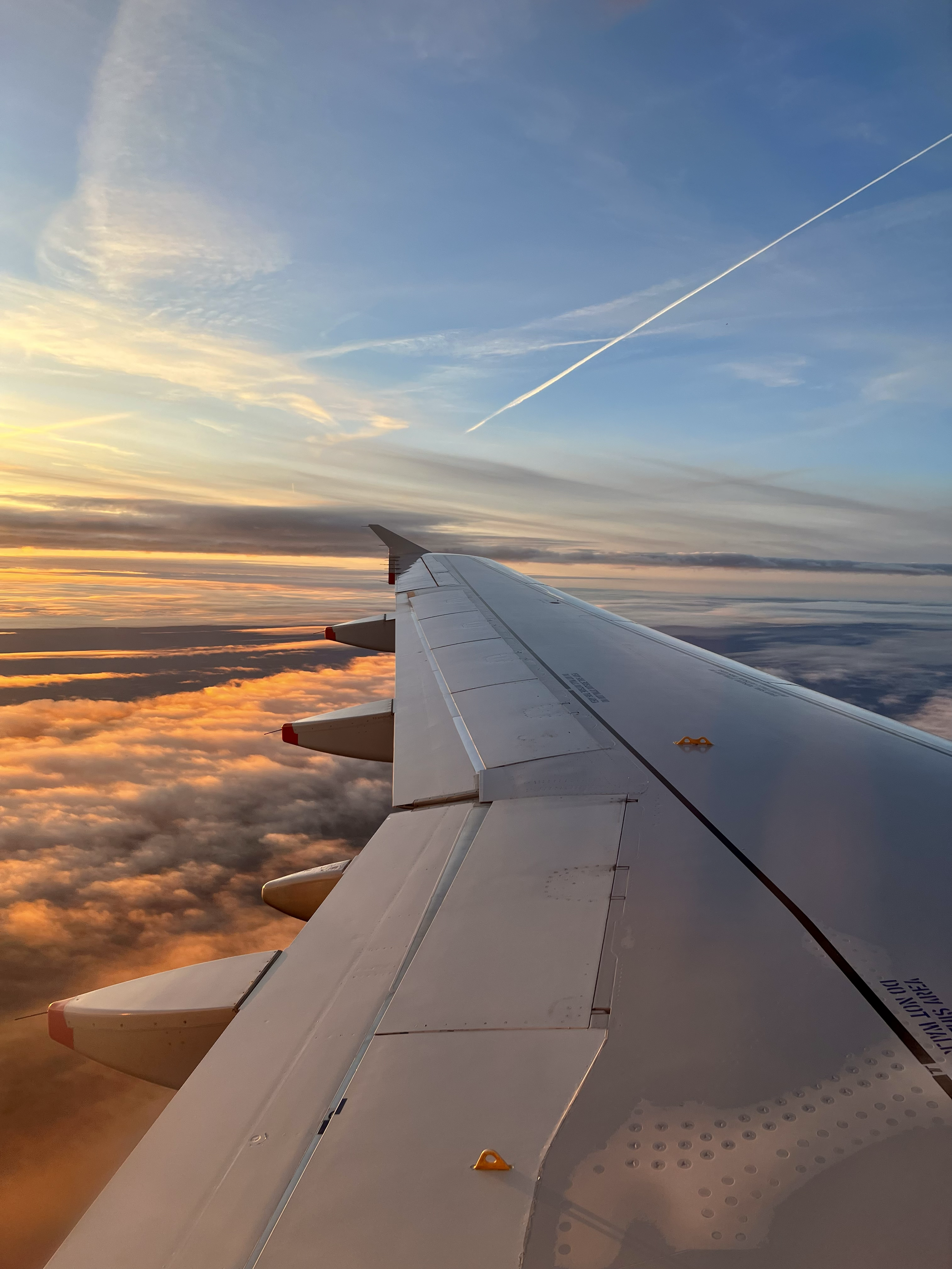 View from an airplane window showing the airplane wing, with a sunset sky and clouds below.