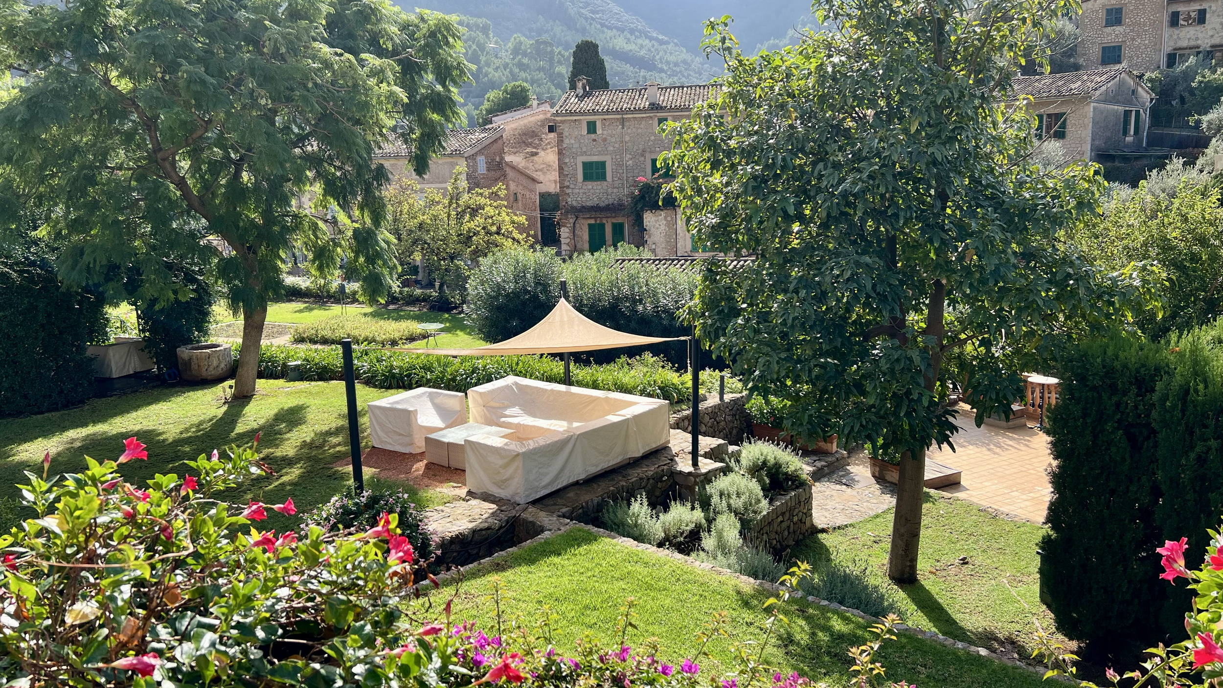 A lush garden with green grass, colorful flowers, tall trees, and stone buildings in the background. There is an outdoor seating area with a white sofa, chairs, and a shaded canopy.