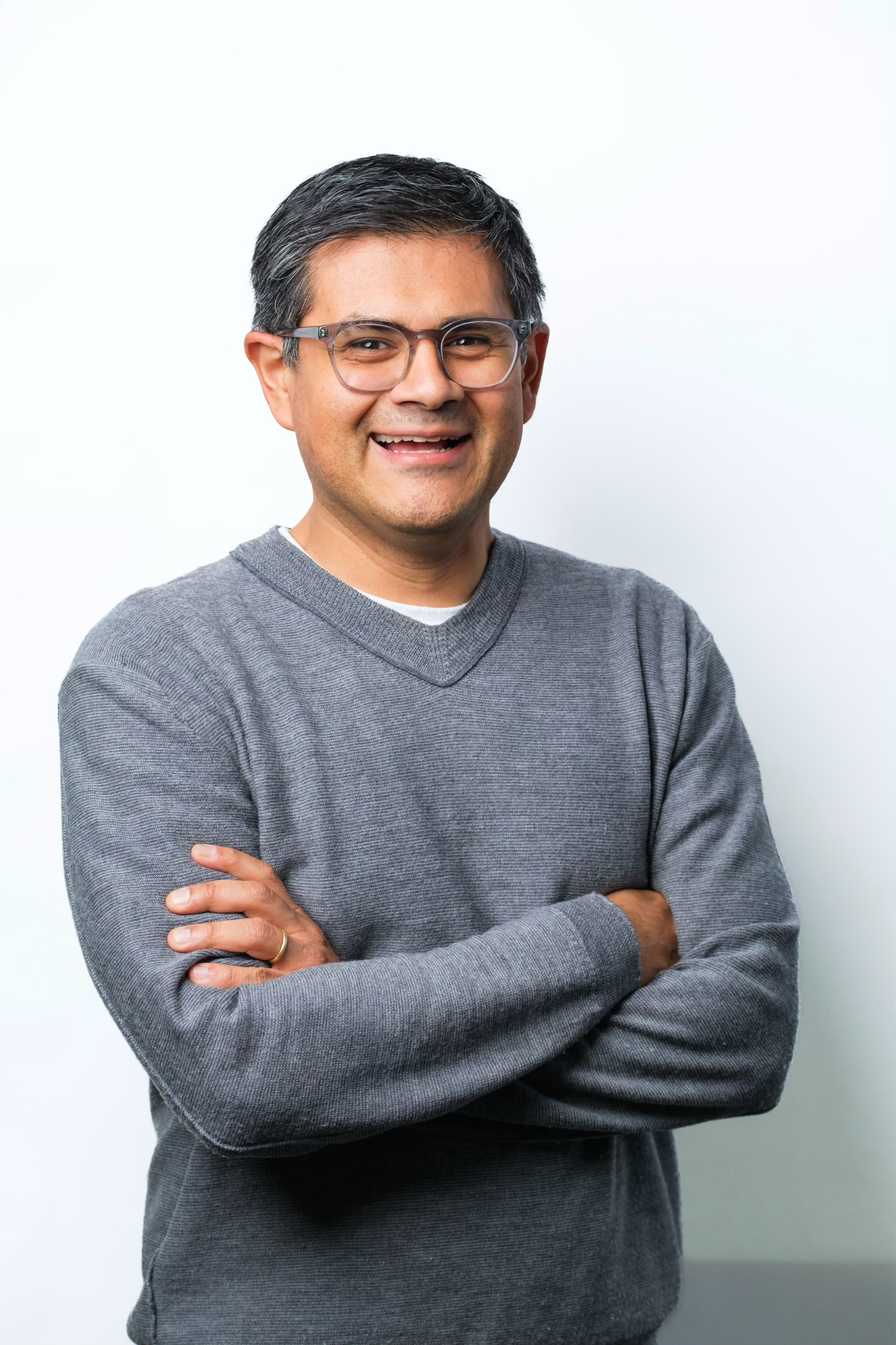 A middle-aged man with short, dark, slightly gray hair and glasses, smiling, with arms crossed, wearing a gray sweater over a white shirt, against a plain white background.