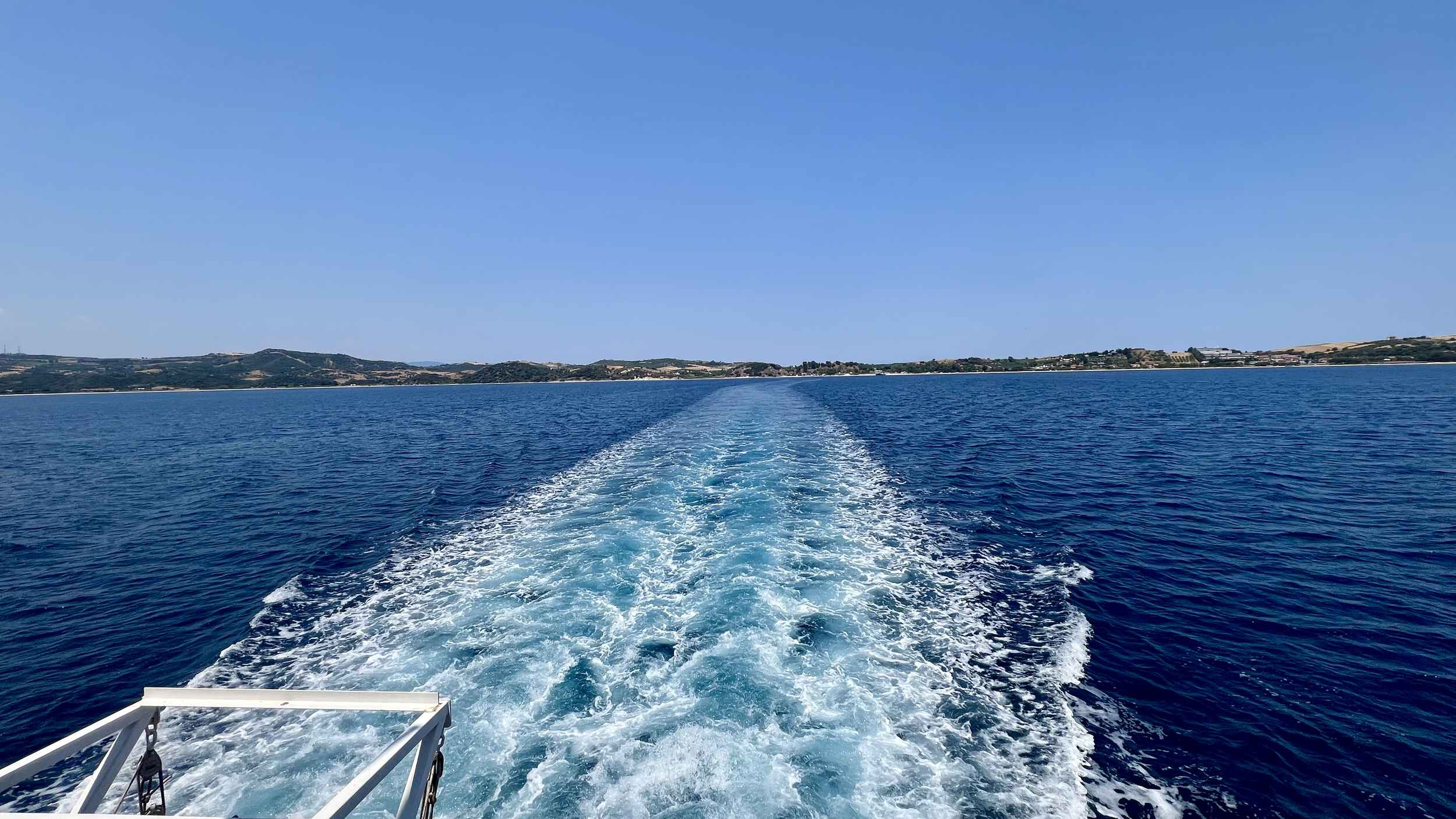 View from a boat showing a blue ocean with a wake trail leading to distant land with hills and some buildings under a clear blue sky.