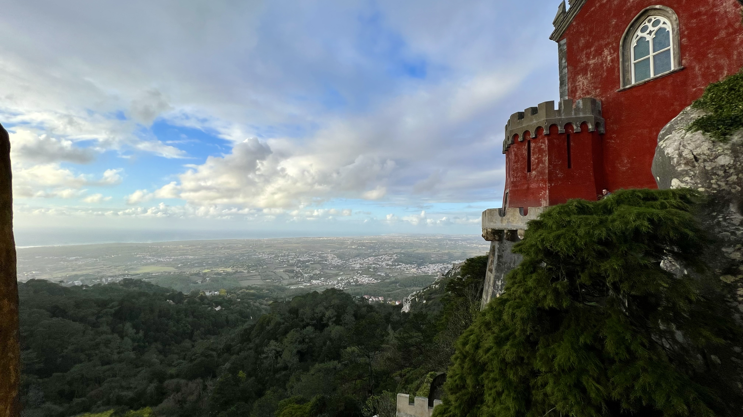 A view from a castle overlooking a vast landscape with forests, fields, and a coastal area under a partly cloudy sky.