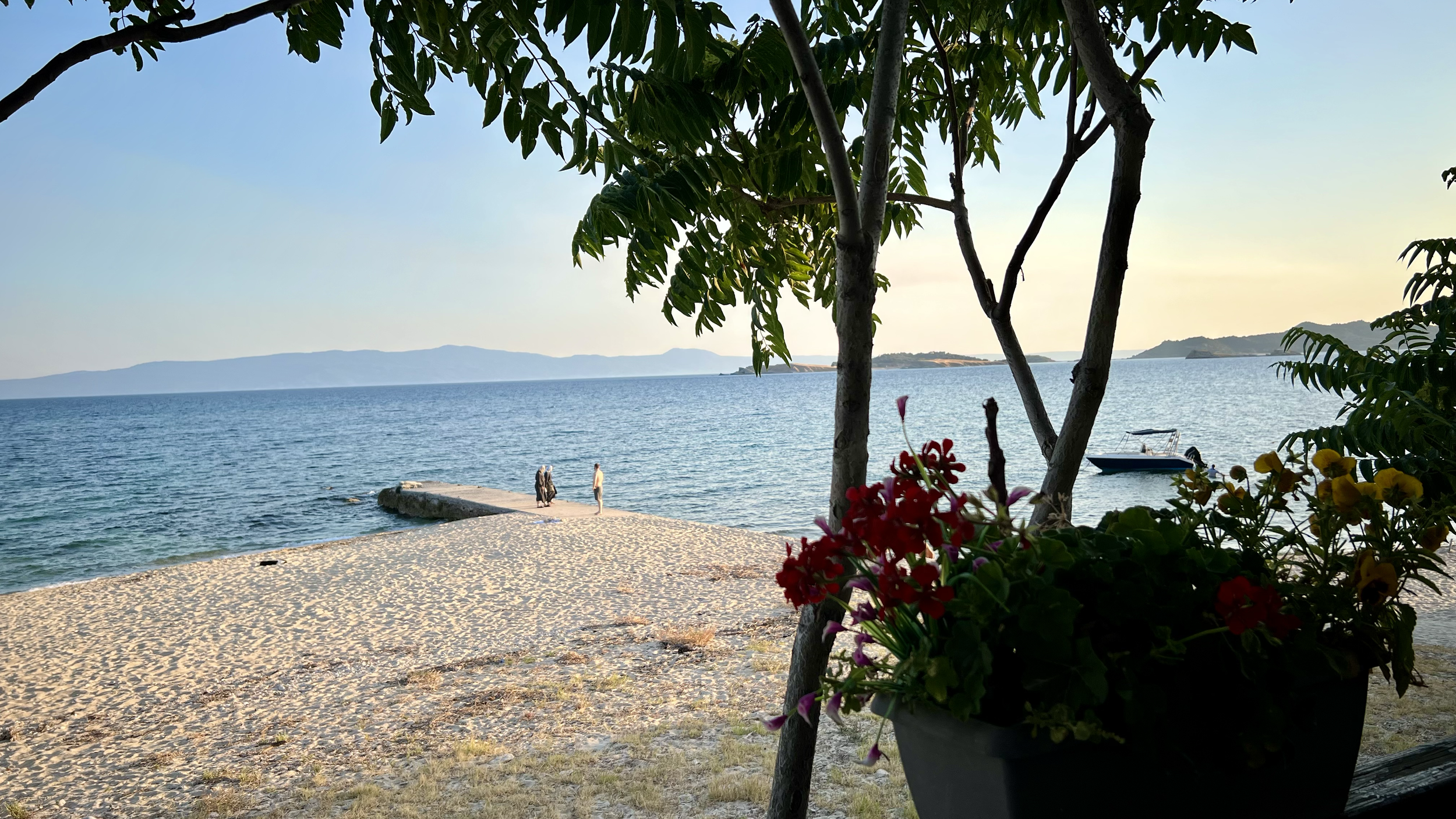 A view of a beach with sand and water, framed by trees, with a boat docked nearby, and a couple walking along the shoreline during sunset.