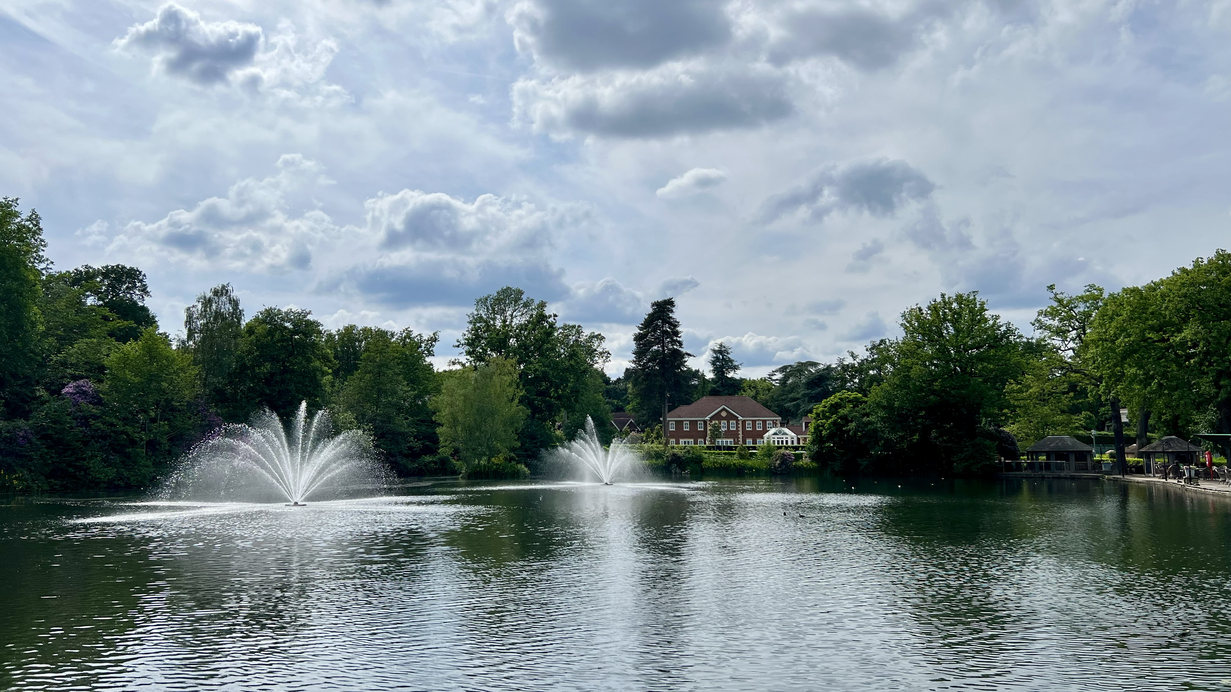 A serene lake scene with two fountains spraying water, surrounded by green trees, a brick house in the background, and a cloudy sky overhead.