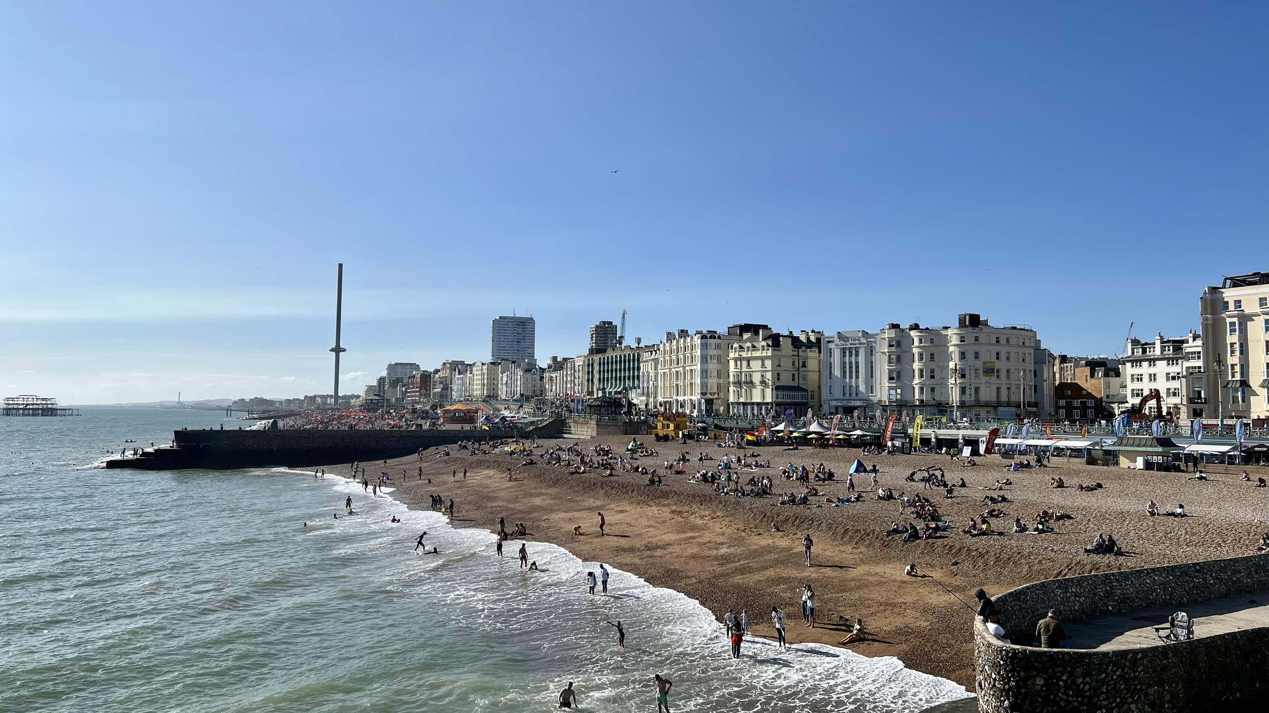 Beach with many people, city buildings in the background, and a body of water in the foreground on a sunny day.