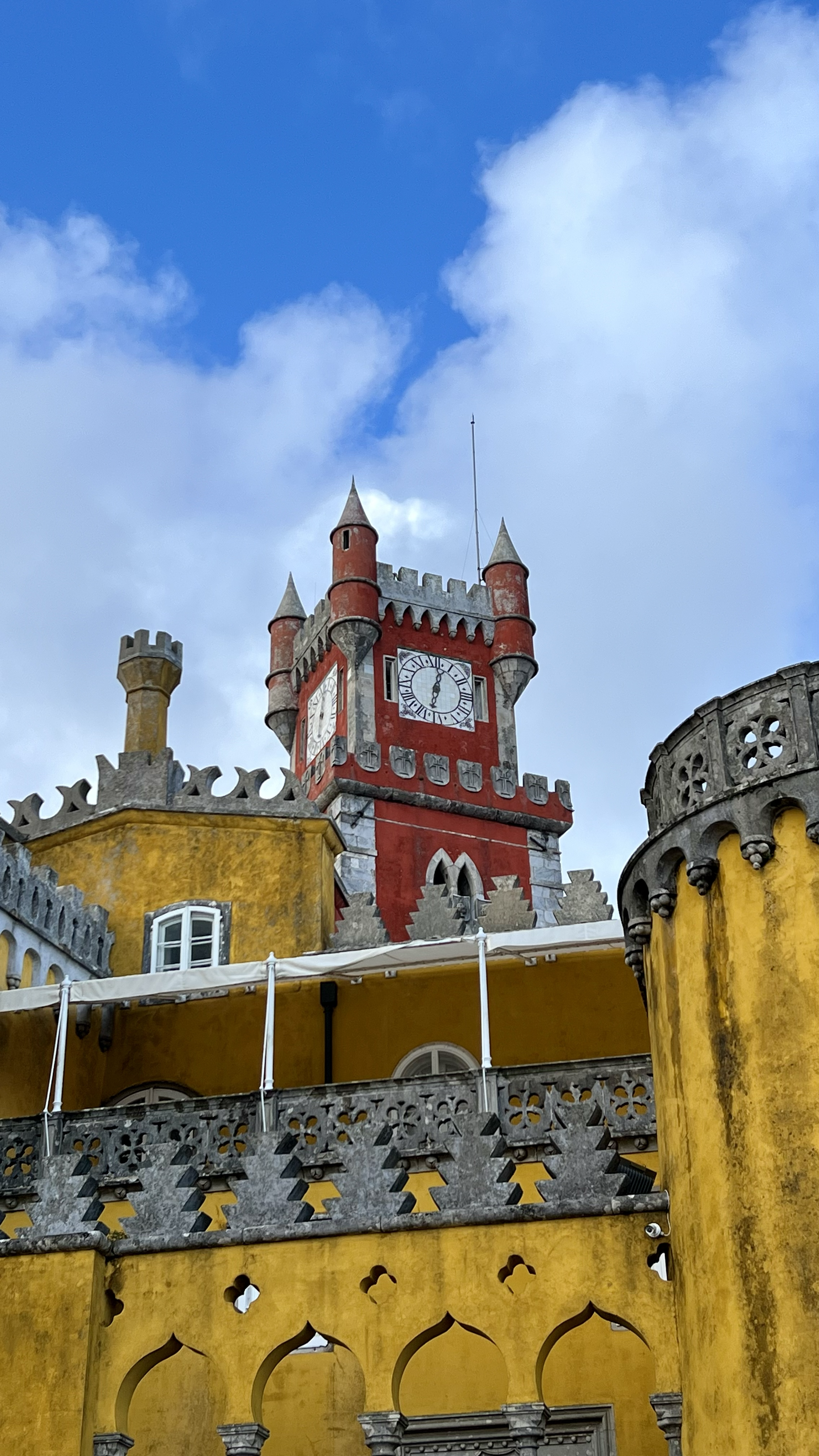 Colorful castle with yellow and red walls, turrets, and a clock tower against a cloudy blue sky.