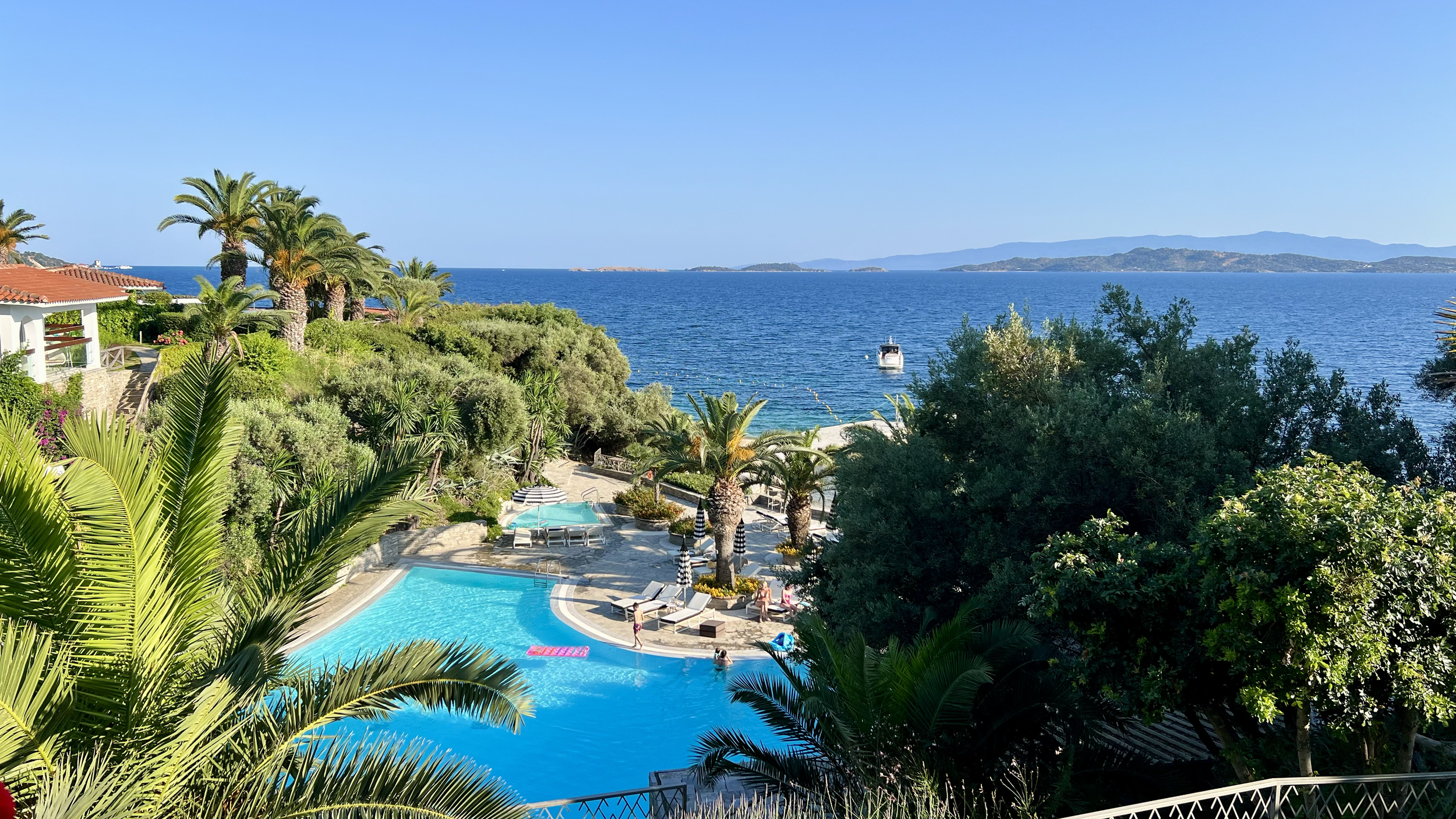 A sunny view of a resort with a swimming pool, surrounded by palm trees and lush greenery, overlooking the ocean with a boat and distant islands.