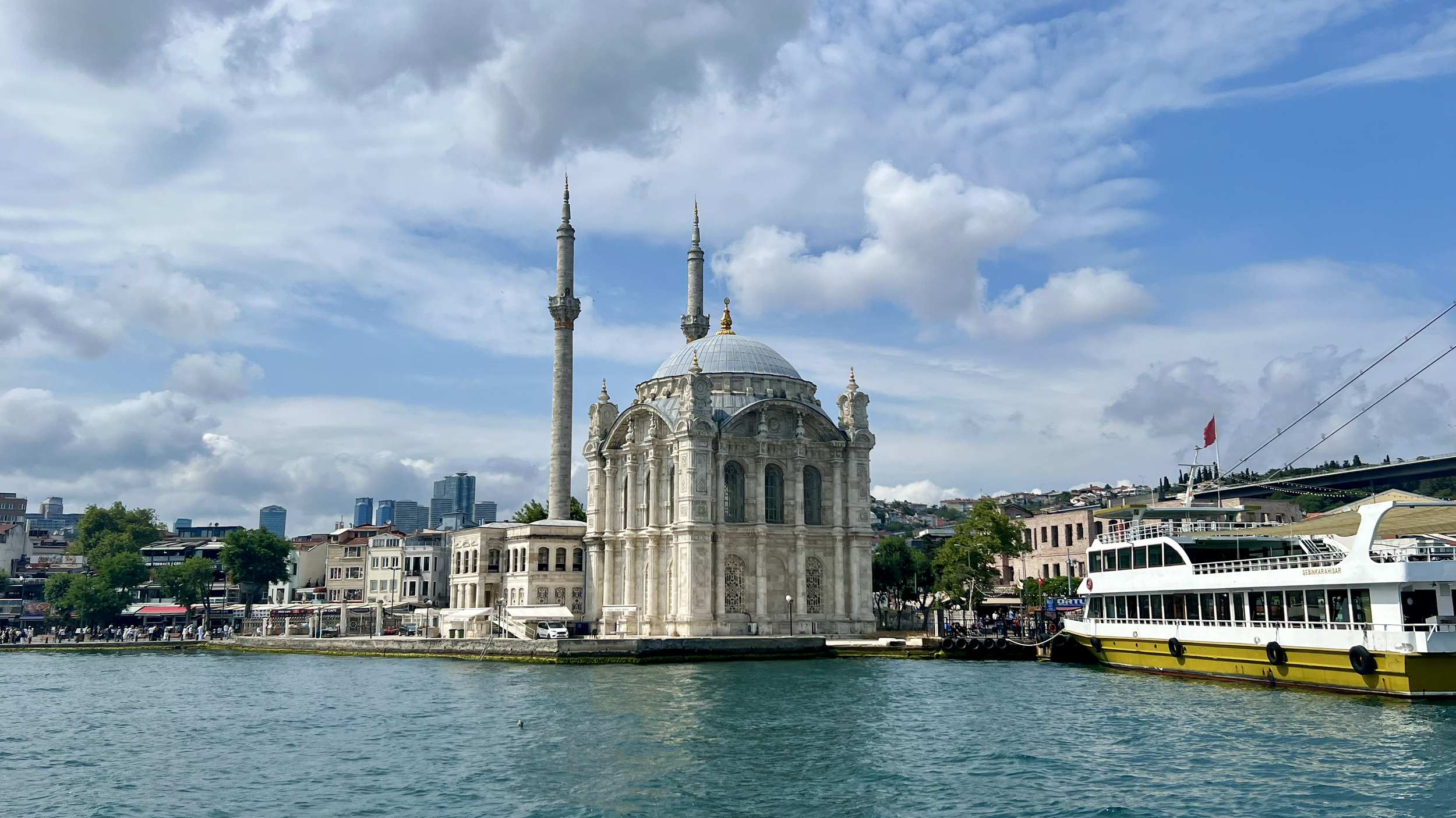 View of the Ortaköy Mosque located by the water in Istanbul, Turkey, with a boat docked nearby and a city skyline in the background.