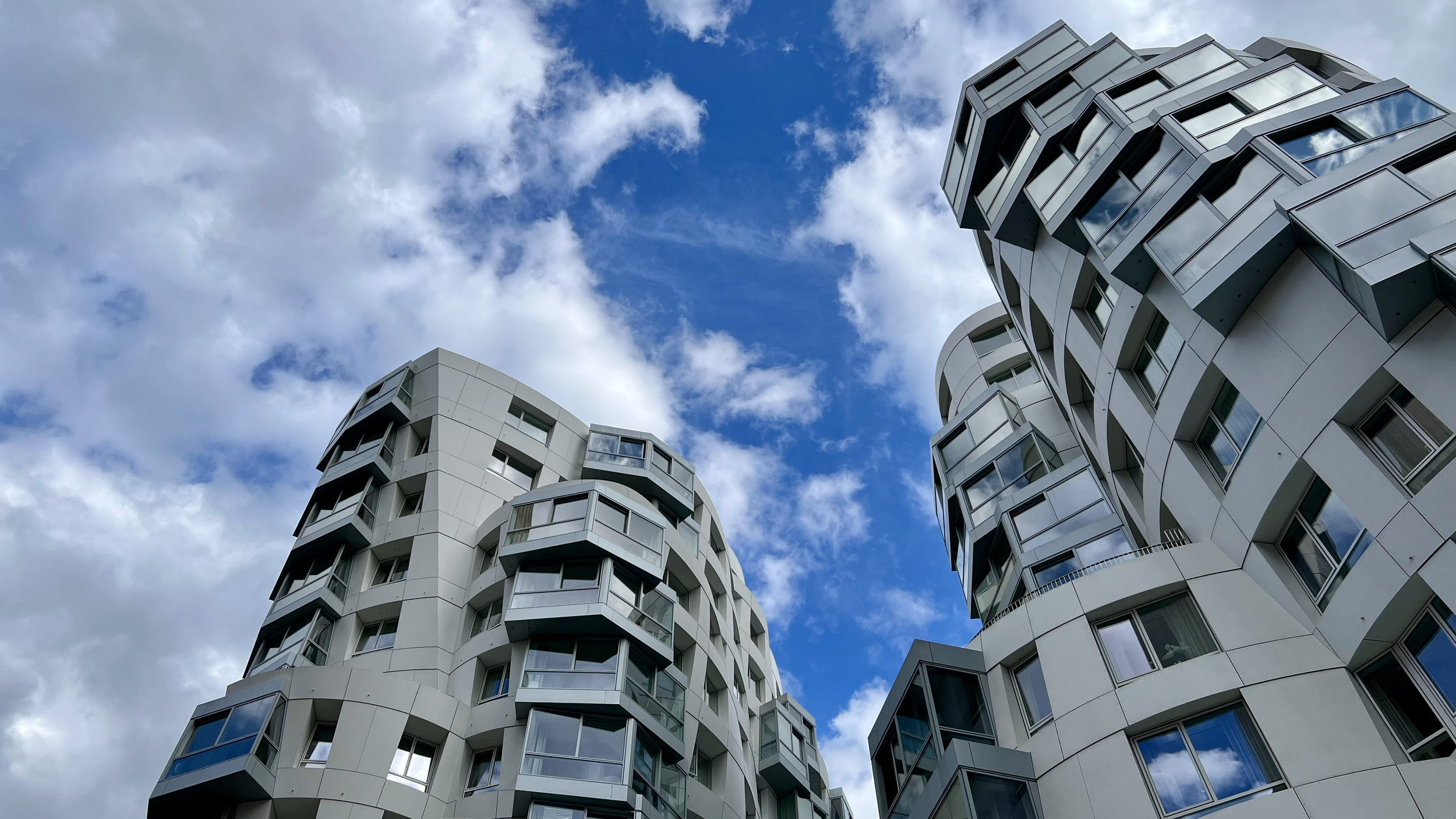 Low-angle view of modern, white apartment buildings with irregular, curved facades and numerous windows, set against a partly cloudy sky.