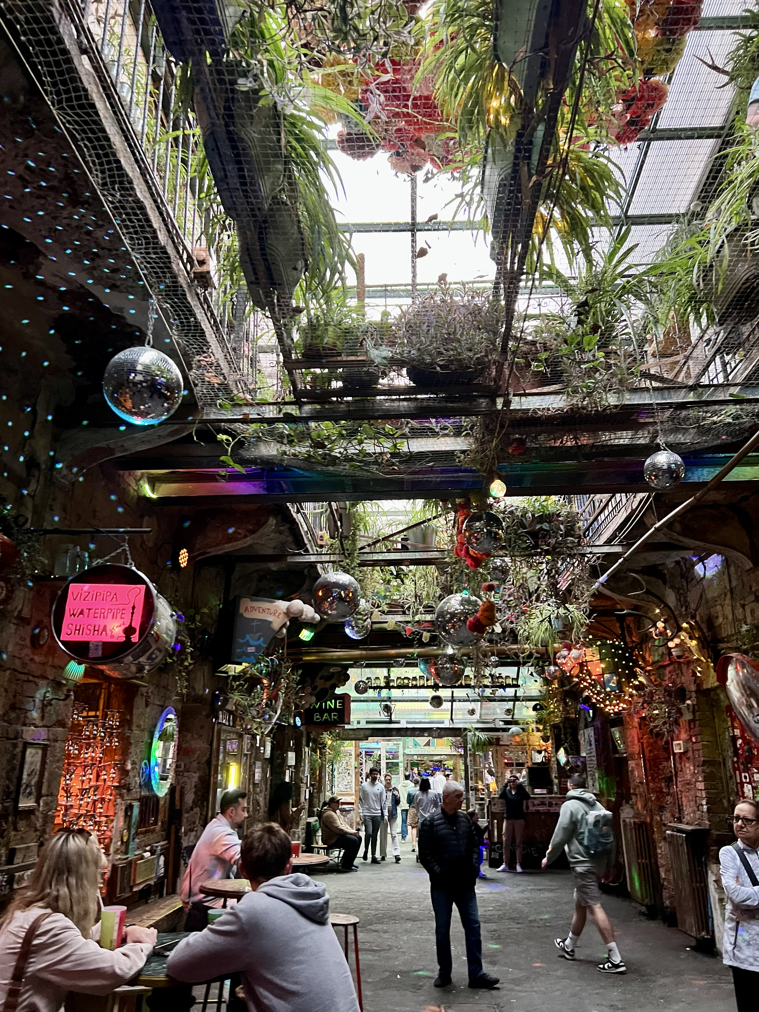 Inside a lively, eclectic bar or restaurant with exposed brick walls, hanging plants, disco balls, colorful lights, and various signs. People are sitting at tables, standing, and walking through the space, which has a vibrant, artistic atmosphere.