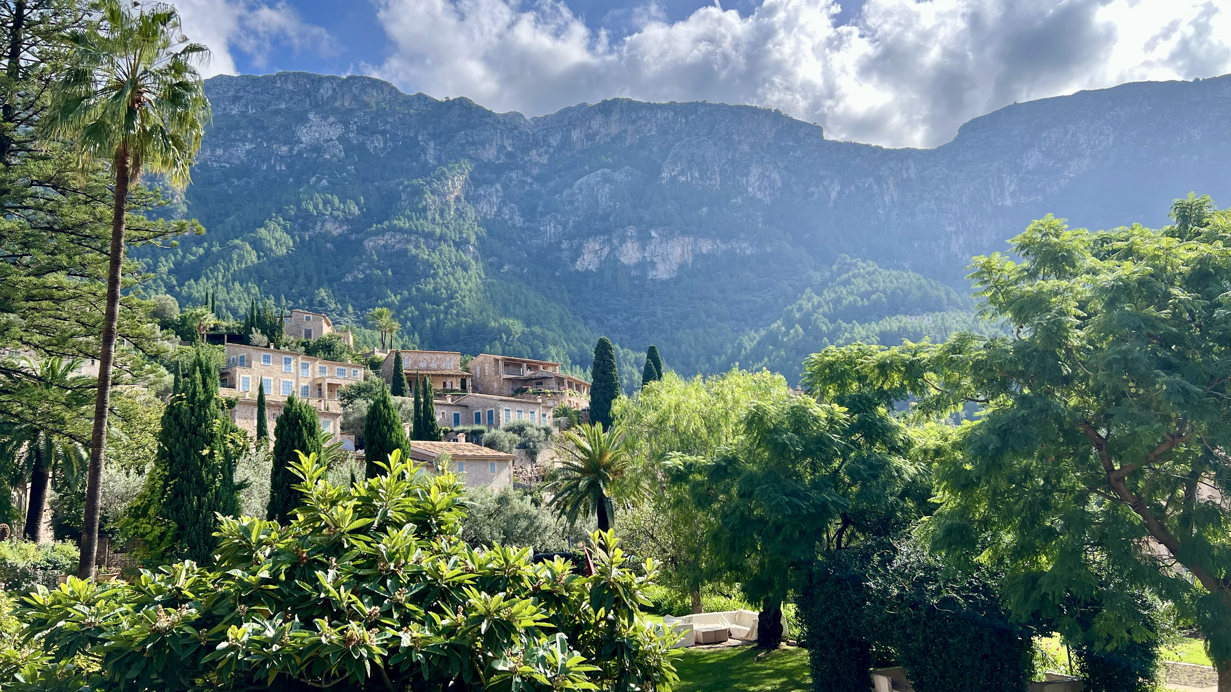 A scenic view of a hillside residential area surrounded by lush greenery, tall trees, and distant mountains under a partly cloudy sky.