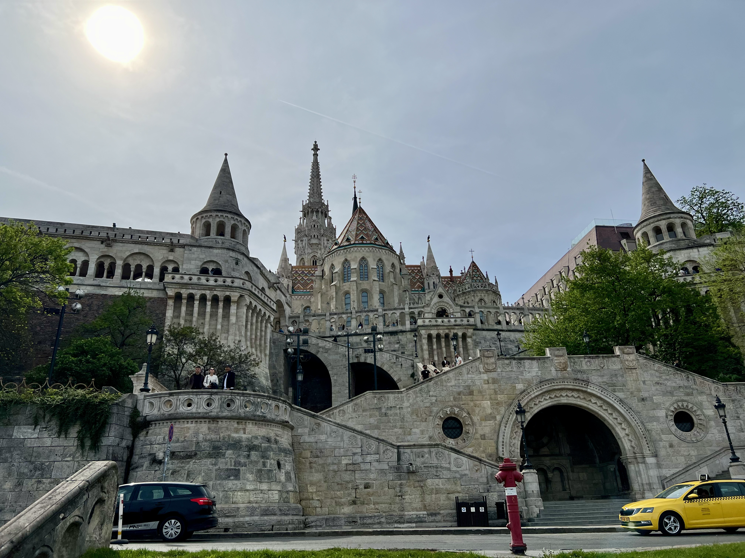 Castle with towers and spires on a hillside, with stairs, trees, and vehicles at the base, during daytime under a cloudy sky.