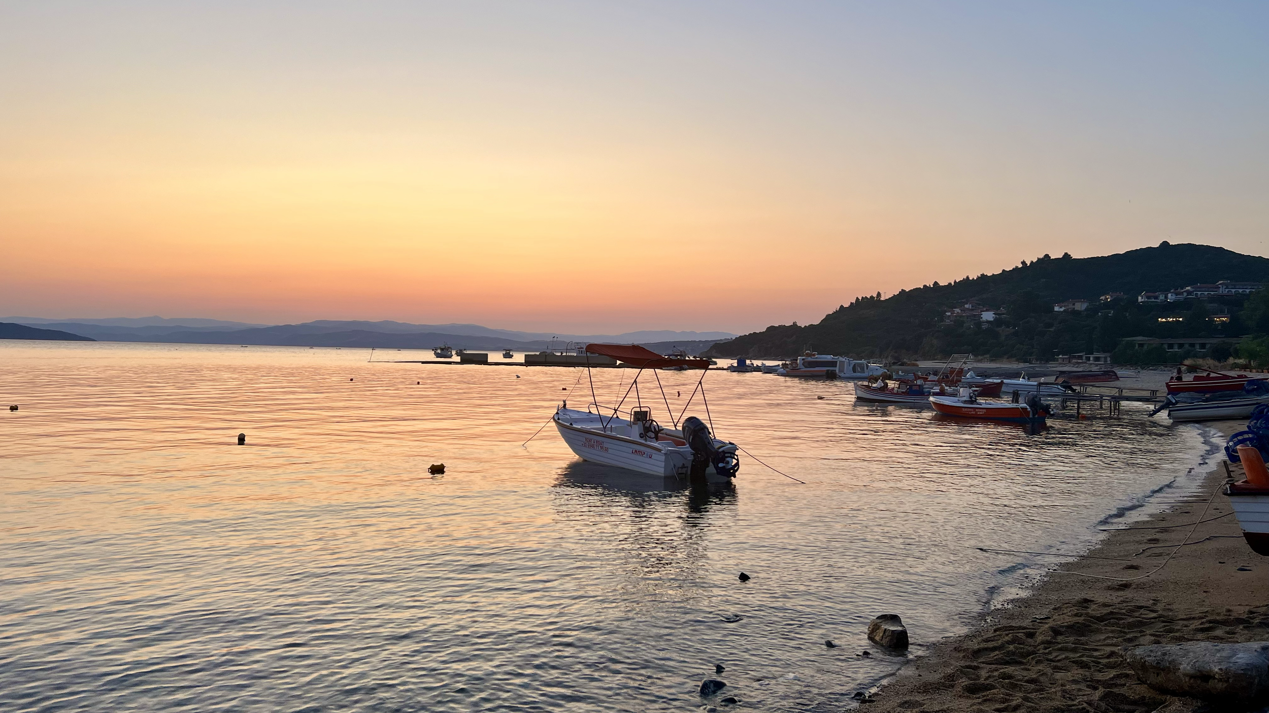Boats anchored near the shoreline of a lake at sunset, with hills in the background and a clear colorful sky.