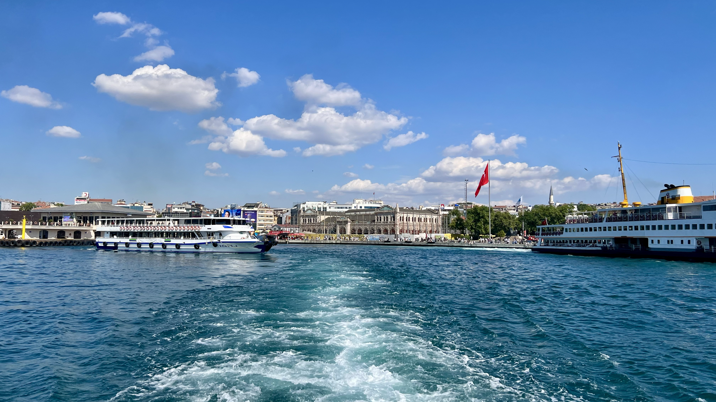 View of boats docked at a harbor with a city skyline in the background under a bright blue sky with scattered clouds.