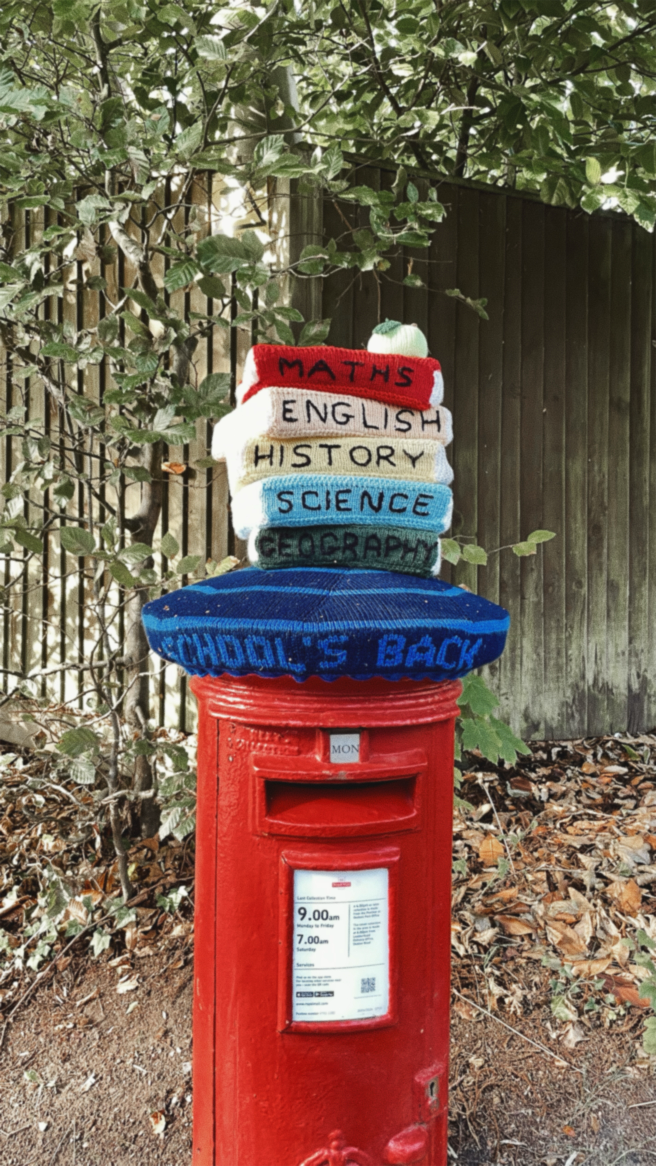 Red postbox decorated with a stack of knitted hats representing school subjects, reading from top to bottom: 'MATHS', 'ENGLISH', 'HISTORY', 'SCIENCE', 'GEOGRAPHY', and 'SCHOOL'S BACK'.