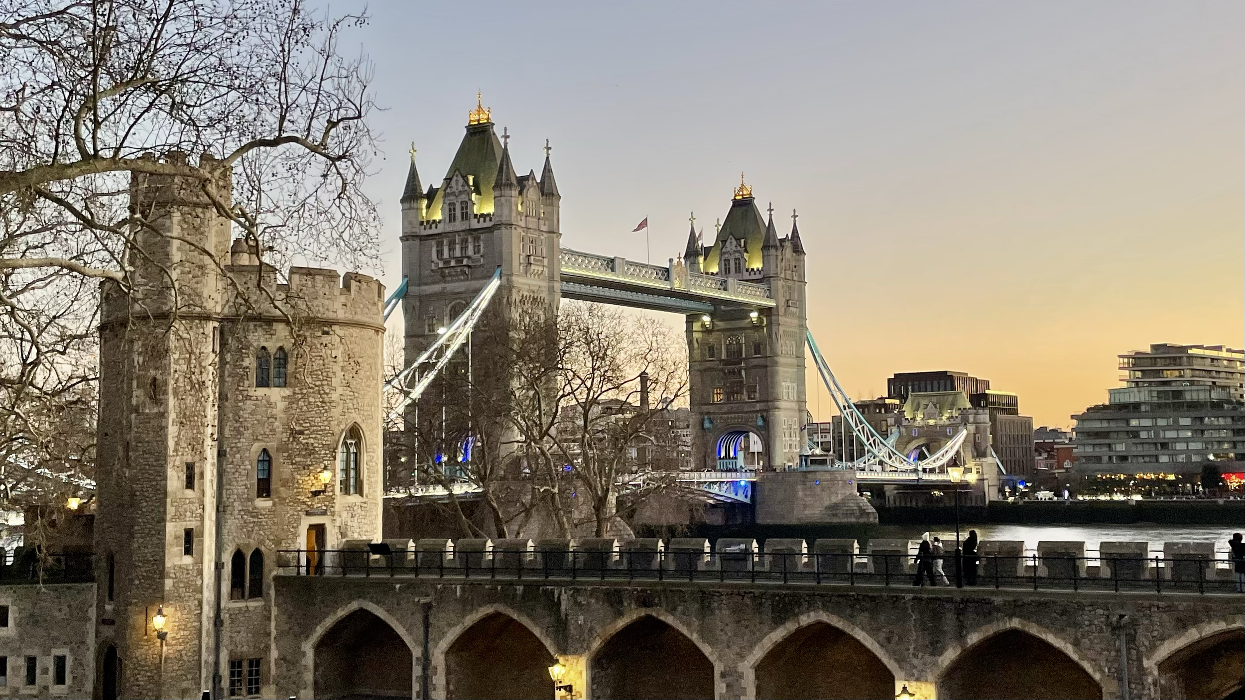 The London Bridge with an old stone tower in the foreground, trees, and modern buildings along the river at sunset.