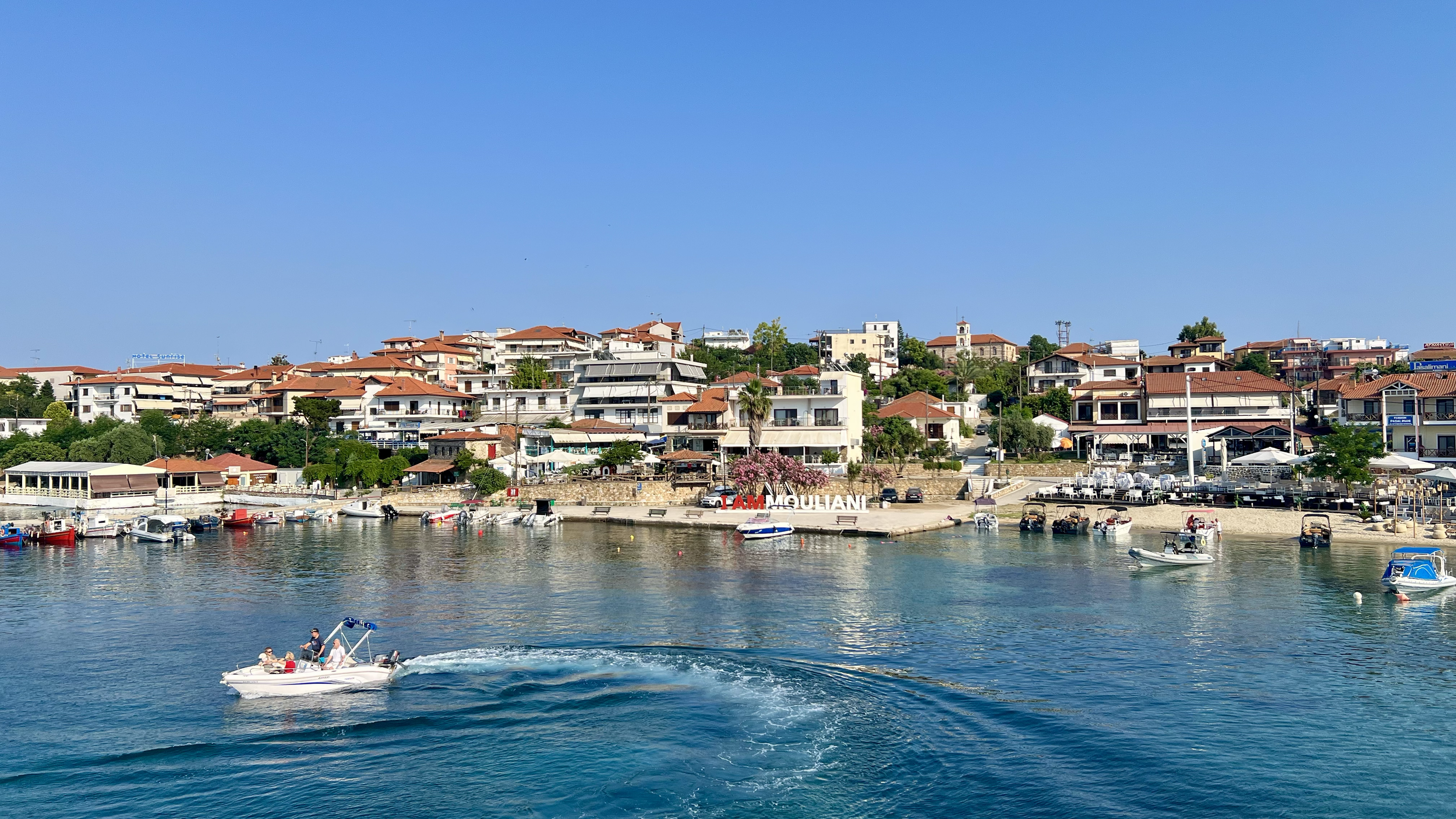A waterfront view of a coastal town with boats in the water and houses on a hillside, under a clear blue sky.