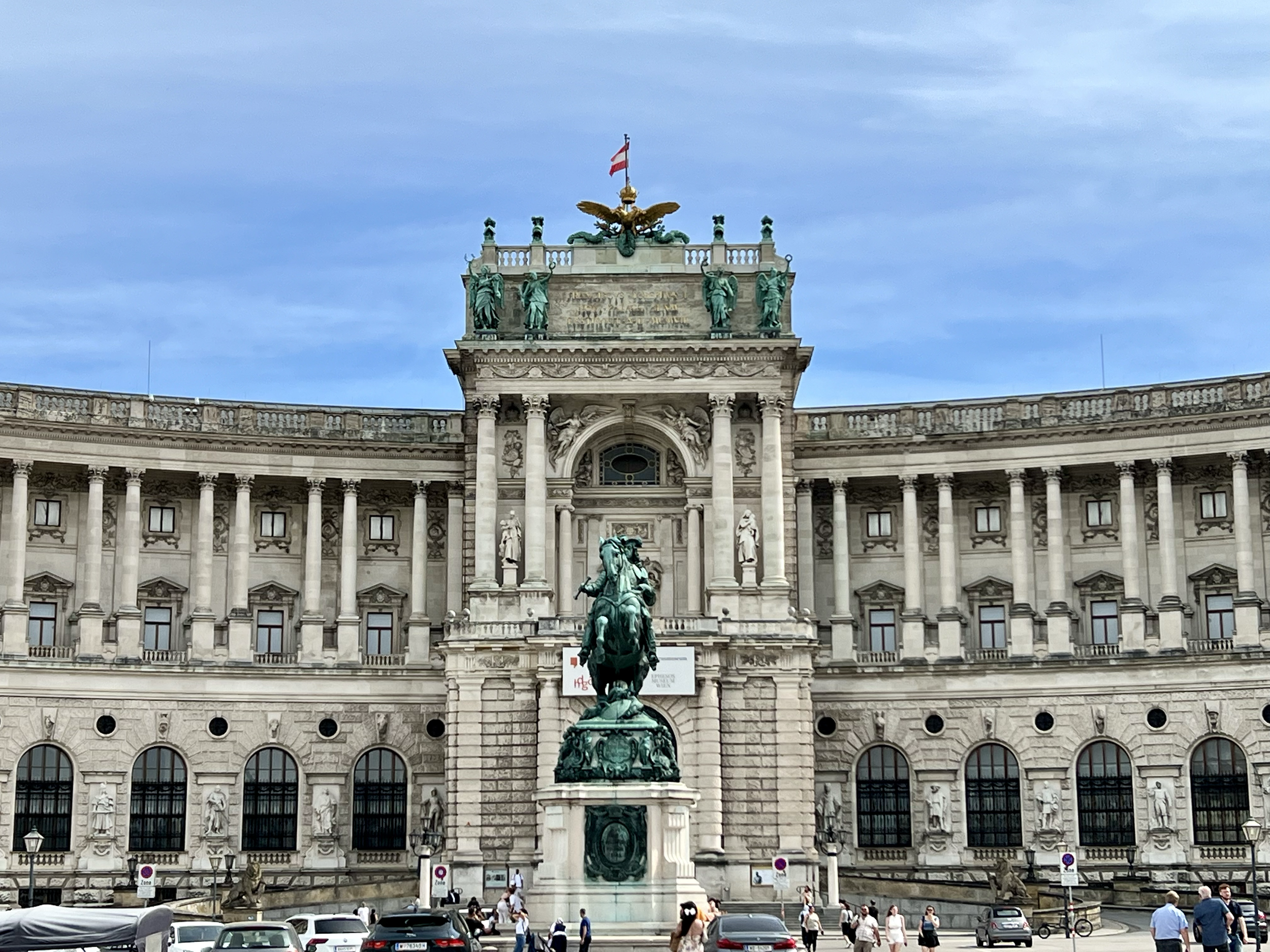 The Hofburg Palace in Vienna, Austria, with a central horse-mounted statue and a large building featuring classical architecture, decorative sculptures, and the Austrian flag on top, under a partly cloudy sky.