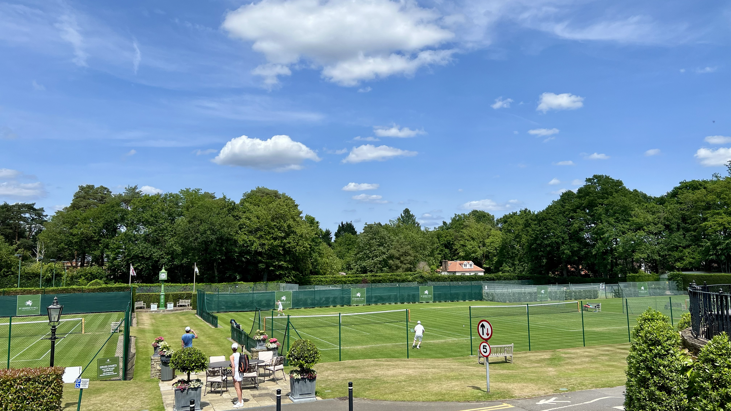 Tennis courts with players, surrounded by green trees and a blue sky with scattered clouds.
