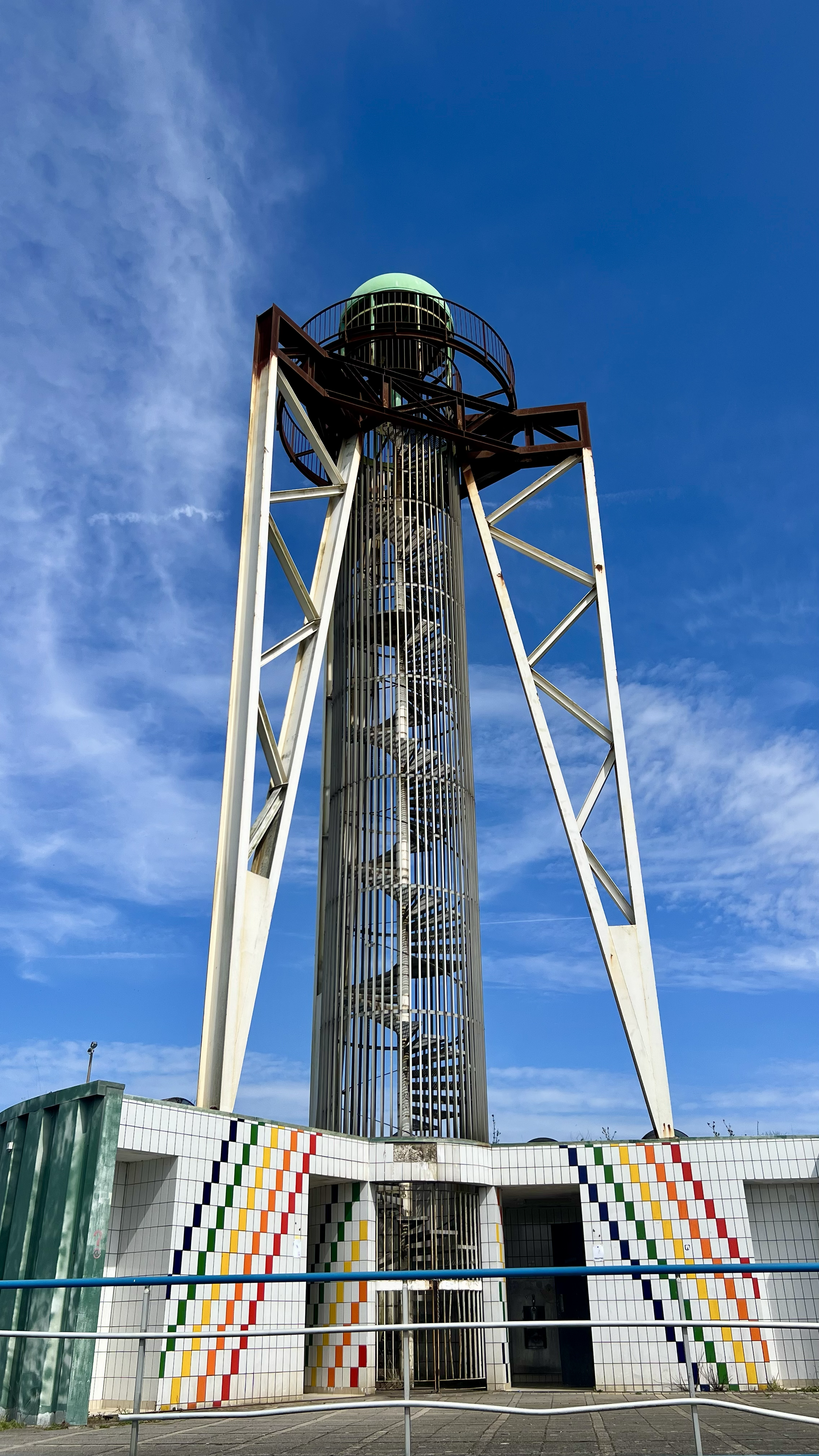 A tall, spiral staircase inside a metal cage tower with a green dome on top, outside against a blue sky.