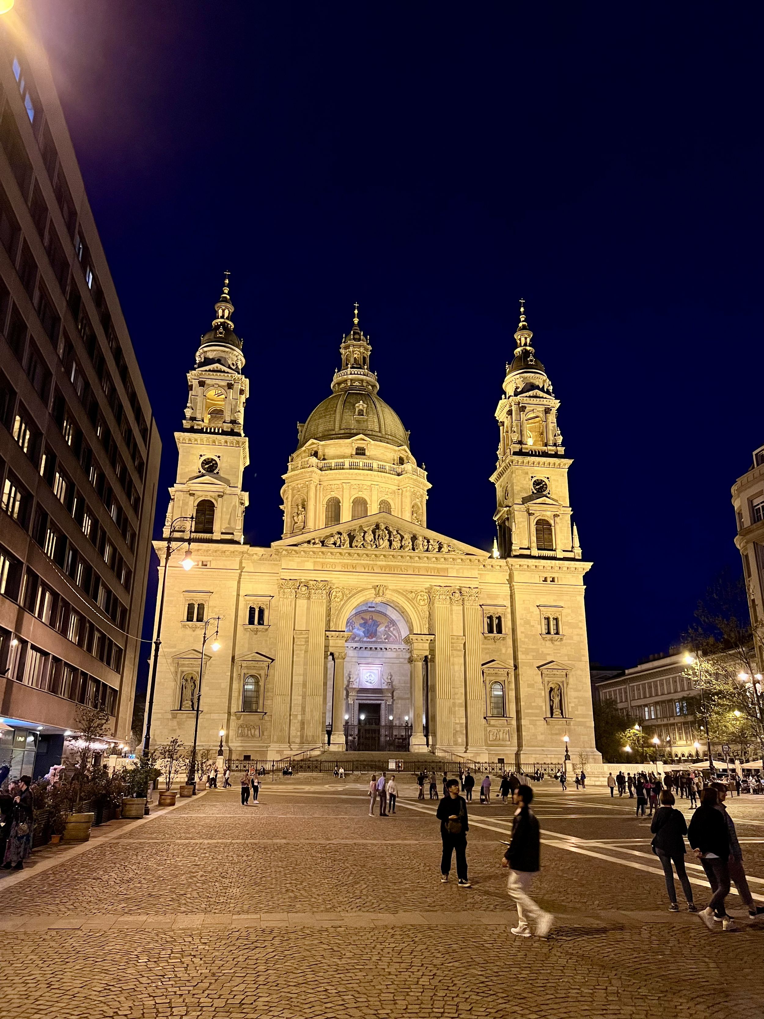 Night view of a large, illuminated historic cathedral with twin towers and a central dome, surrounded by a cobblestone plaza with people walking and sitting.