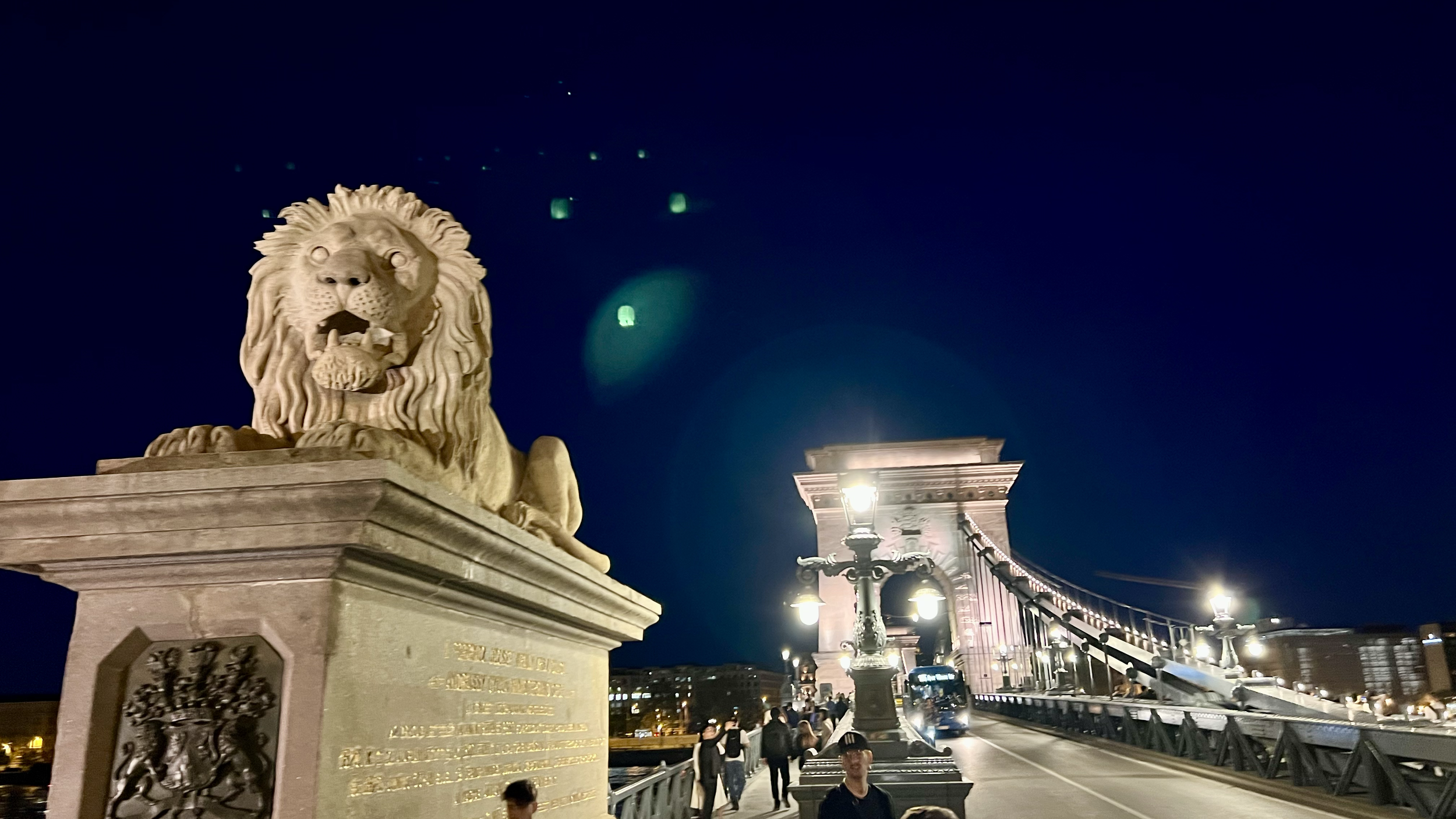 Nighttime view of the Chain Bridge in Budapest with a stone lion statue in the foreground. The bridge is illuminated with lights, and people are walking across it.