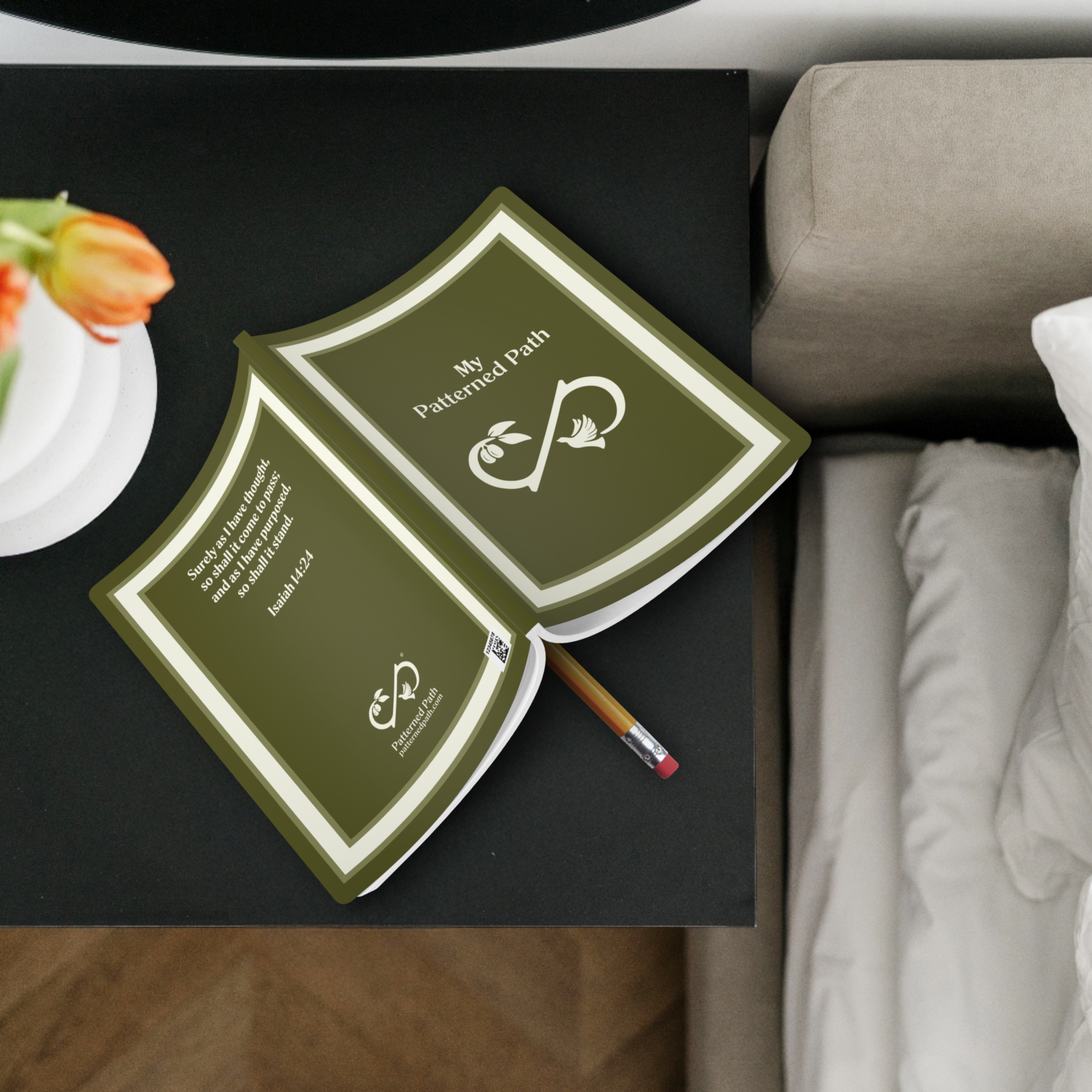 Olive Green Journal with cream square border. Text "My Patterned Path" in center. Infinity symbol with olive branch and dove. Back cover shows scripture. On end table next to bed.