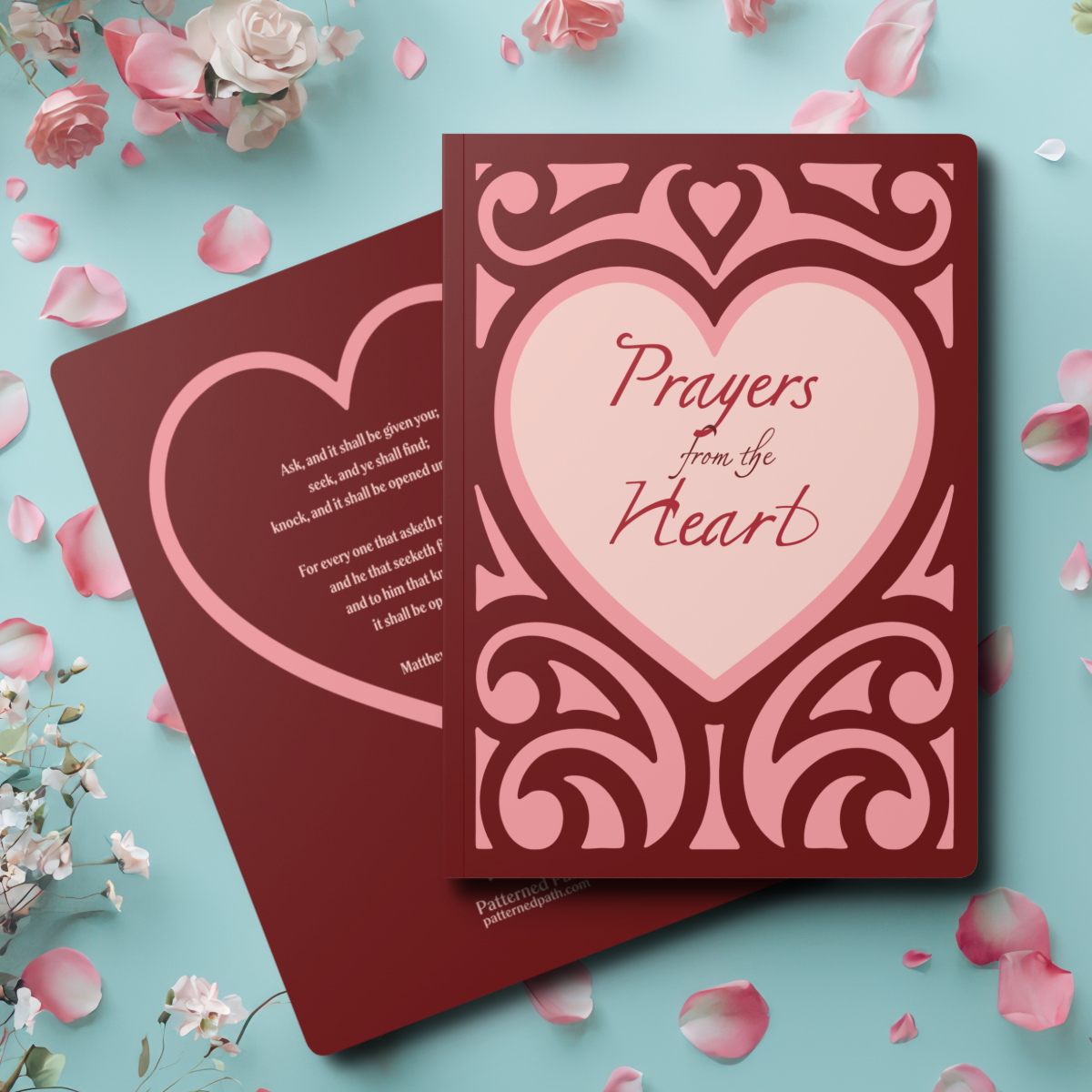 Two Journals on top of one another. top journal shows front cover (Burgendy with pink ornate scroll work with heart in center , text "Prayers from the Heart" in center. Bottom journal shows pink heart with scripture. Laying on a teal blue table