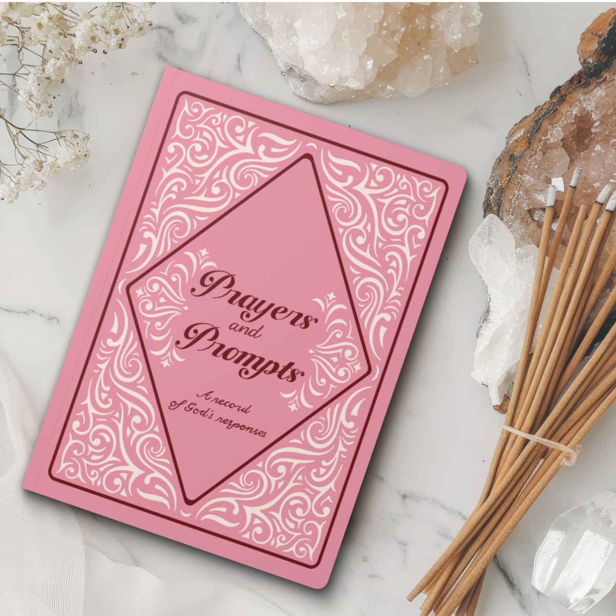 Pink Journal with cream ornate scroll work surrounding text "Prayers and Prompts, A record of God's responses" laying on a marble slab with dried flowers and cotton with incense next to it.