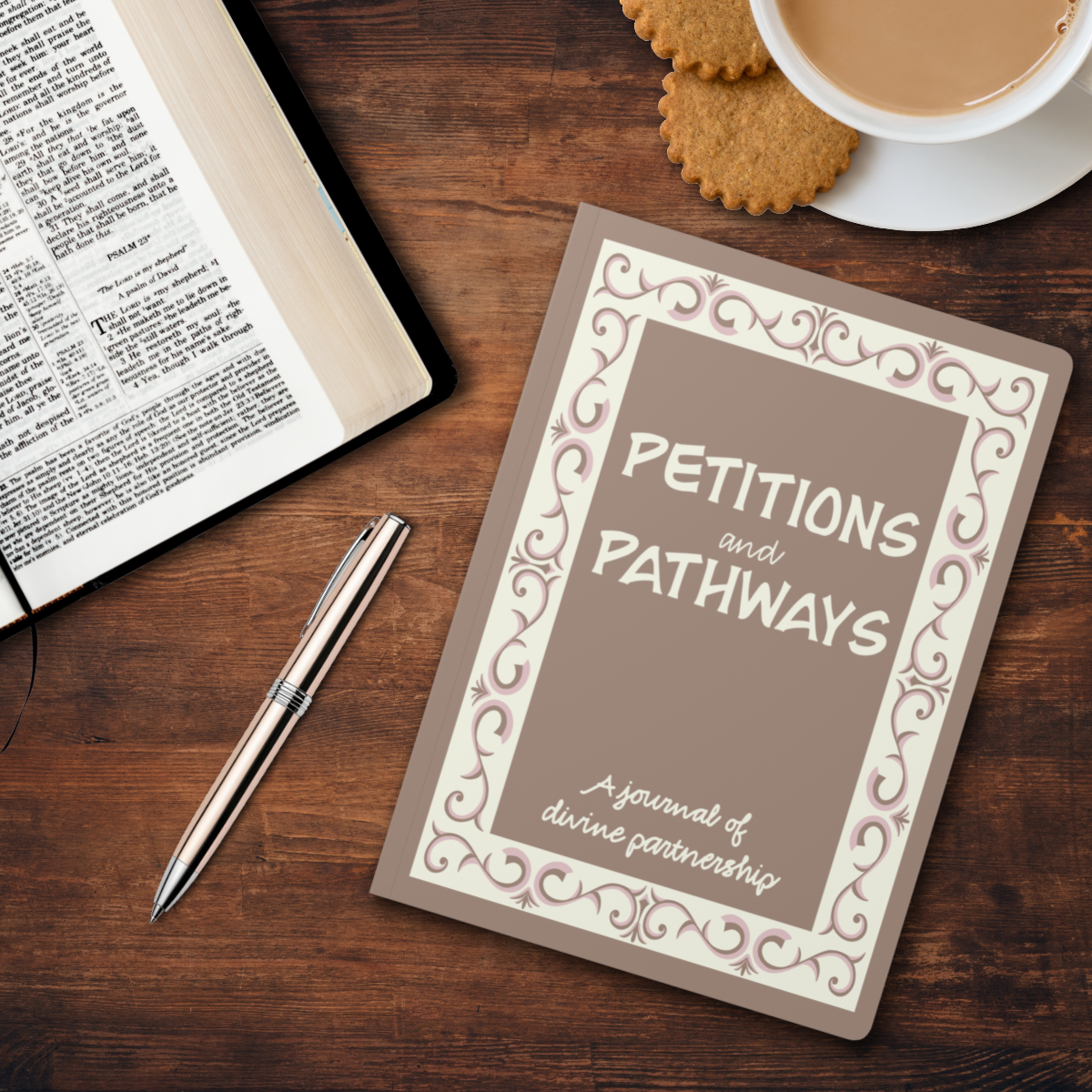 Light brown journal with text "Petitions and Pathways, A journal of divine partnership" written on journal. Cream rectangular border with pink and light brown ornate scroll work within cream border. Pen laying beside journal with scriptures on a desk