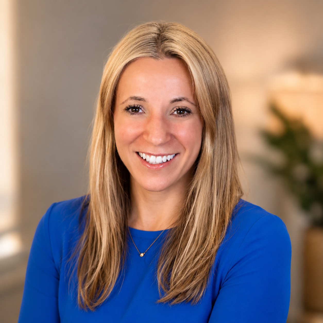 A smiling woman with long blonde hair wearing a blue top and a gold necklace, indoors with a neutral background and blurred plant in the background.