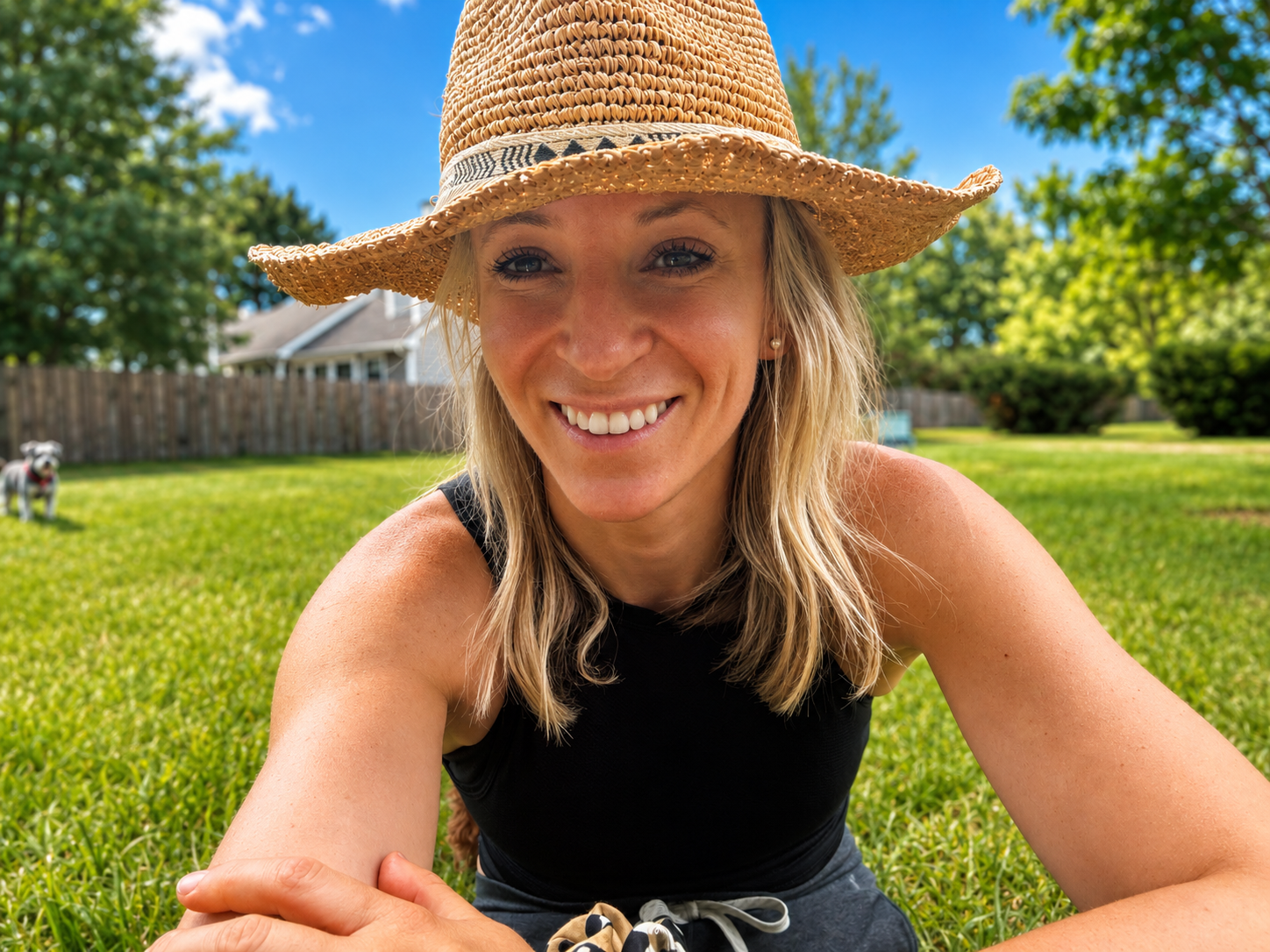A woman with blonde hair, smiling, wearing a wide-brimmed straw hat and a sleeveless black top, lying or sitting on green grass in a backyard with a wooden fence, trees, and a house in the background on a sunny day.