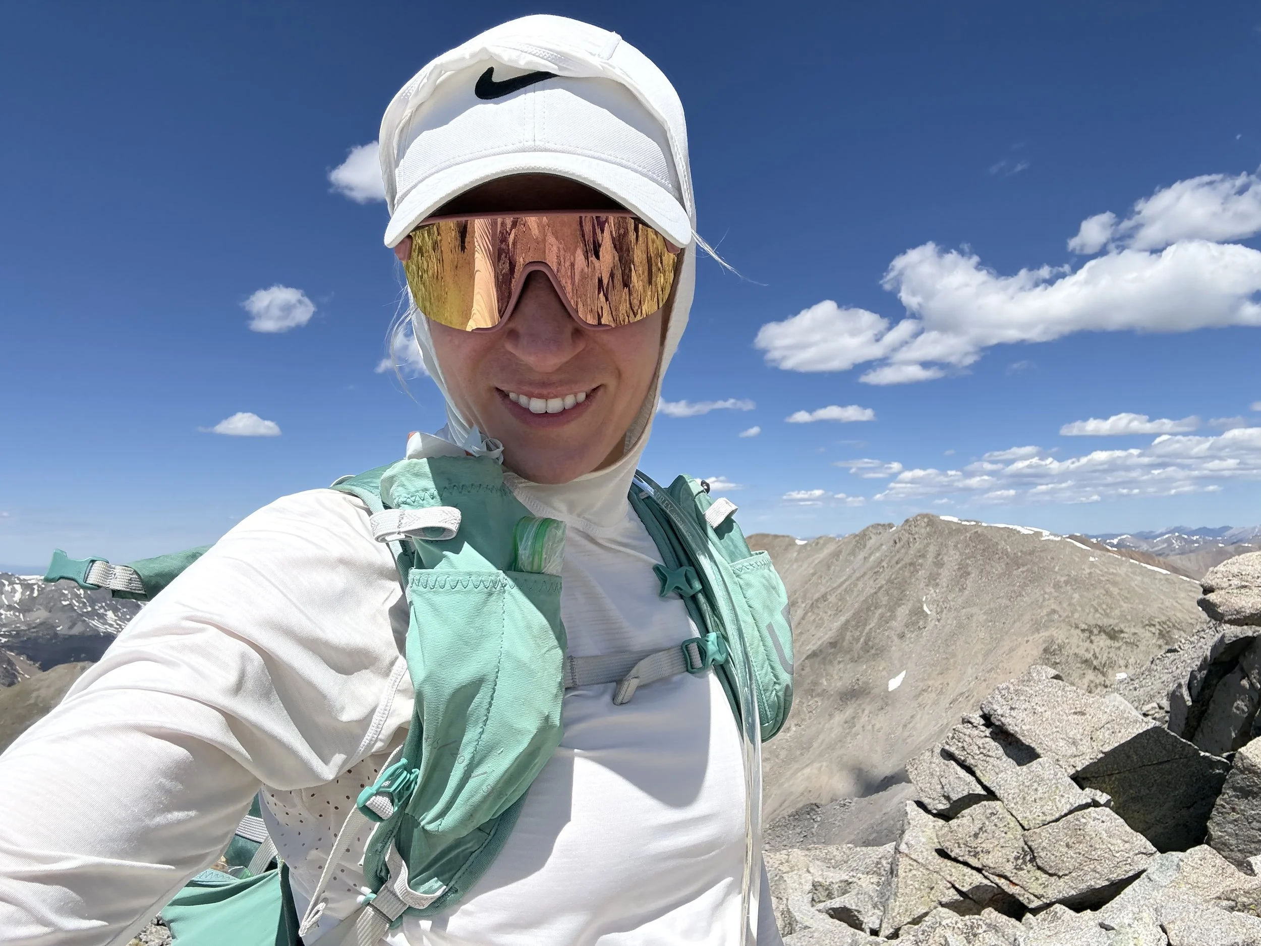 A woman smiling outdoors in a mountainous area, wearing a white cap, reflective sunglasses, and outdoor gear.