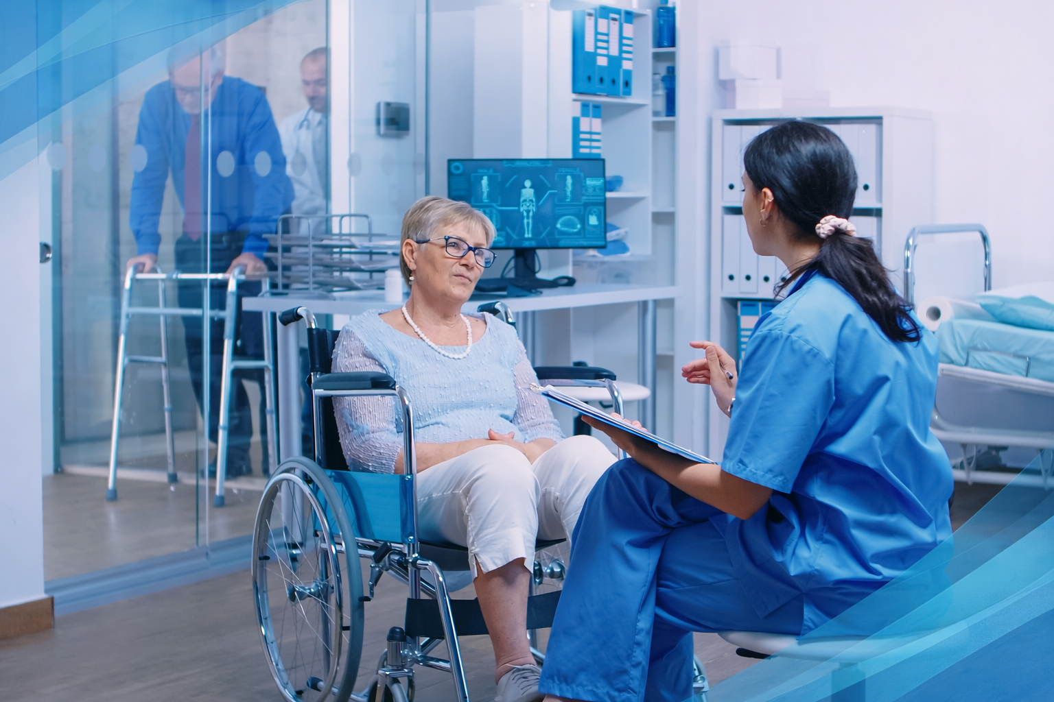 A female nurse in blue scrubs talks to an elderly woman in a wheelchair in a hospital room, with medical equipment and a computer screen displaying a body scan in the background.