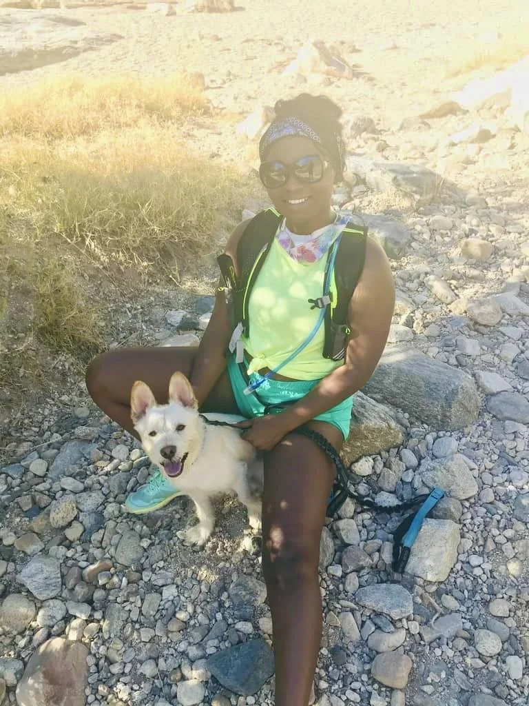 Person sitting on rocky terrain with a white dog, wearing a backpack and sunglasses.