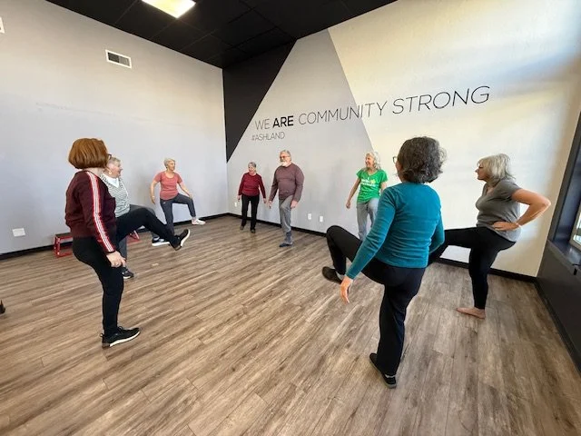 A group of seven women participating in a fitness or exercise class in a room with wooden floors and a large wall with motivational words. The women are stretching or warming up, with some using the wall or chairs for support.