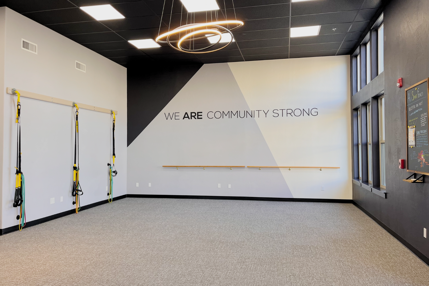 An indoor fitness space with black ceiling tiles, a modern ring-shaped light fixture, and a wall with the message 'WE ARE COMMUNITY STRONG'. The room features vertical wooden handrails and resistance bands hanging on a wall. Large windows are on one 