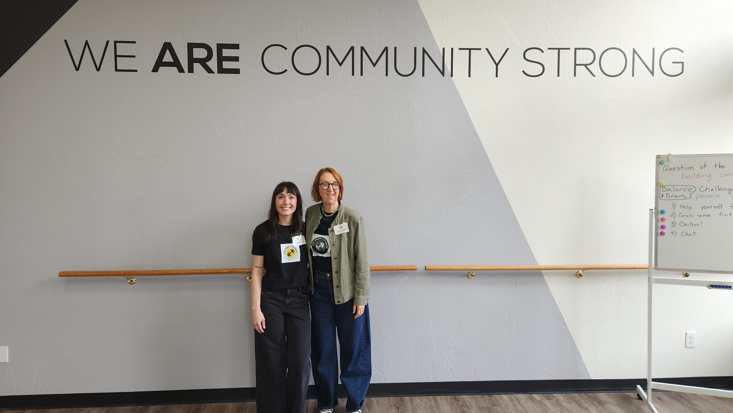 Two women standing side by side in front of a wall with a large sign that reads, "WE ARE COMMUNITY STRONG." One woman has black hair and is wearing a black T-shirt and black pants, while the other woman has red hair and is wearing glasses, a green ja