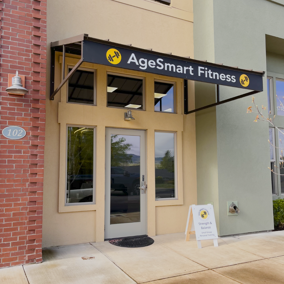 Exterior of a fitness studio named AgeSmart Fitness with a black sign featuring a yellow dumbbell icon. A white sandwich board sign on the sidewalk advertises small group personal training focusing on strength and balance.