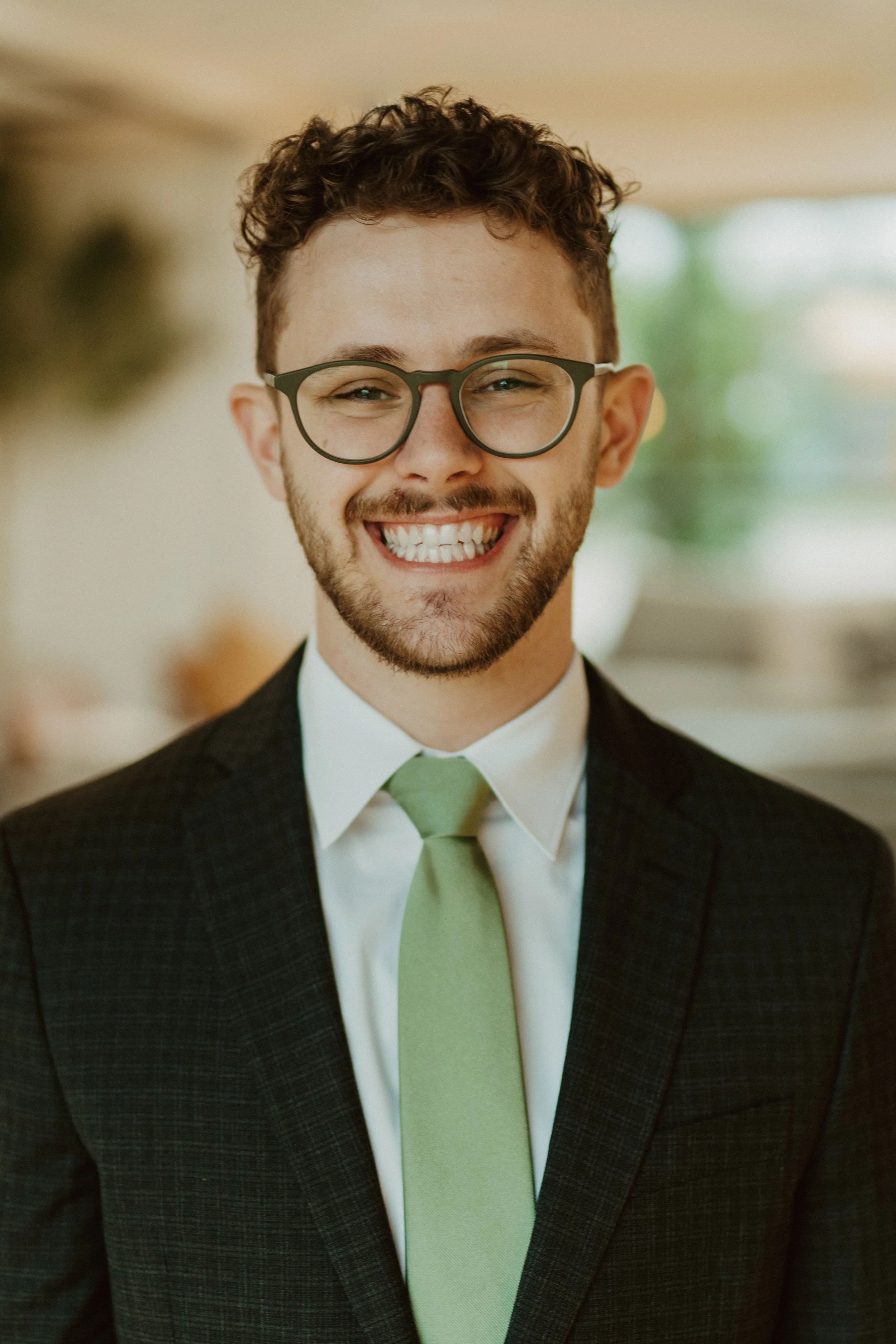 Smiling person wearing glasses, a dark suit, white shirt, and green tie.