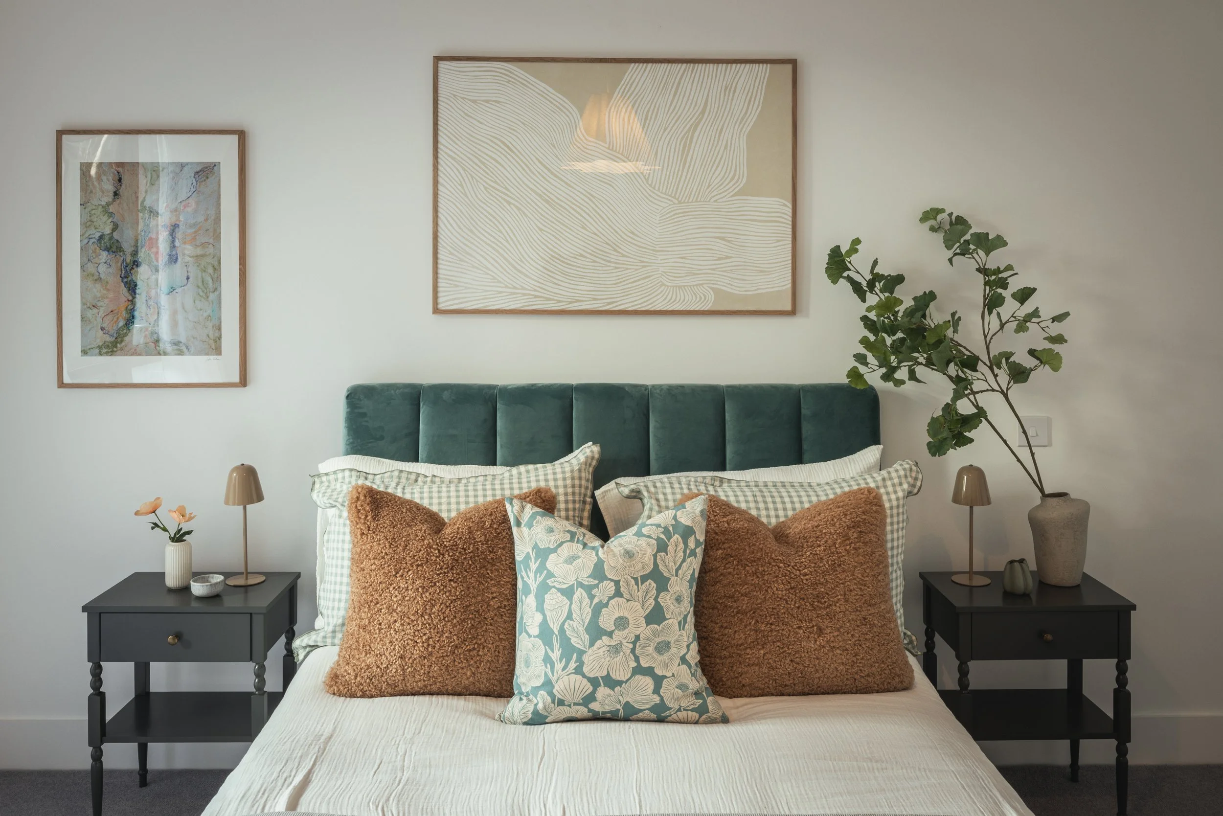A bedroom with a bed, multiple pillows, and two black nightstands. The wall above the bed has framed artwork and a potted plant on the right nightstand. The room has a minimalist decor with a neutral color palette.