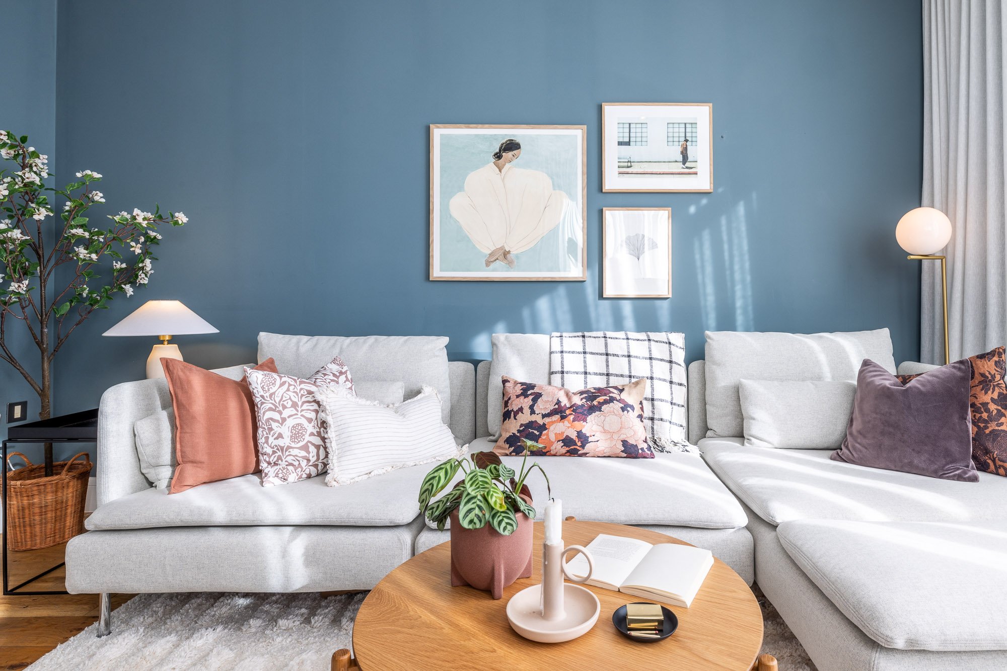 A living room with a light gray sectional sofa decorated with various patterned cushions, a white table lamp, several framed artworks on a blue wall, and a wooden coffee table with decorative items including a potted plant, an open book, and candle holders. Curtains are visible on the right side.