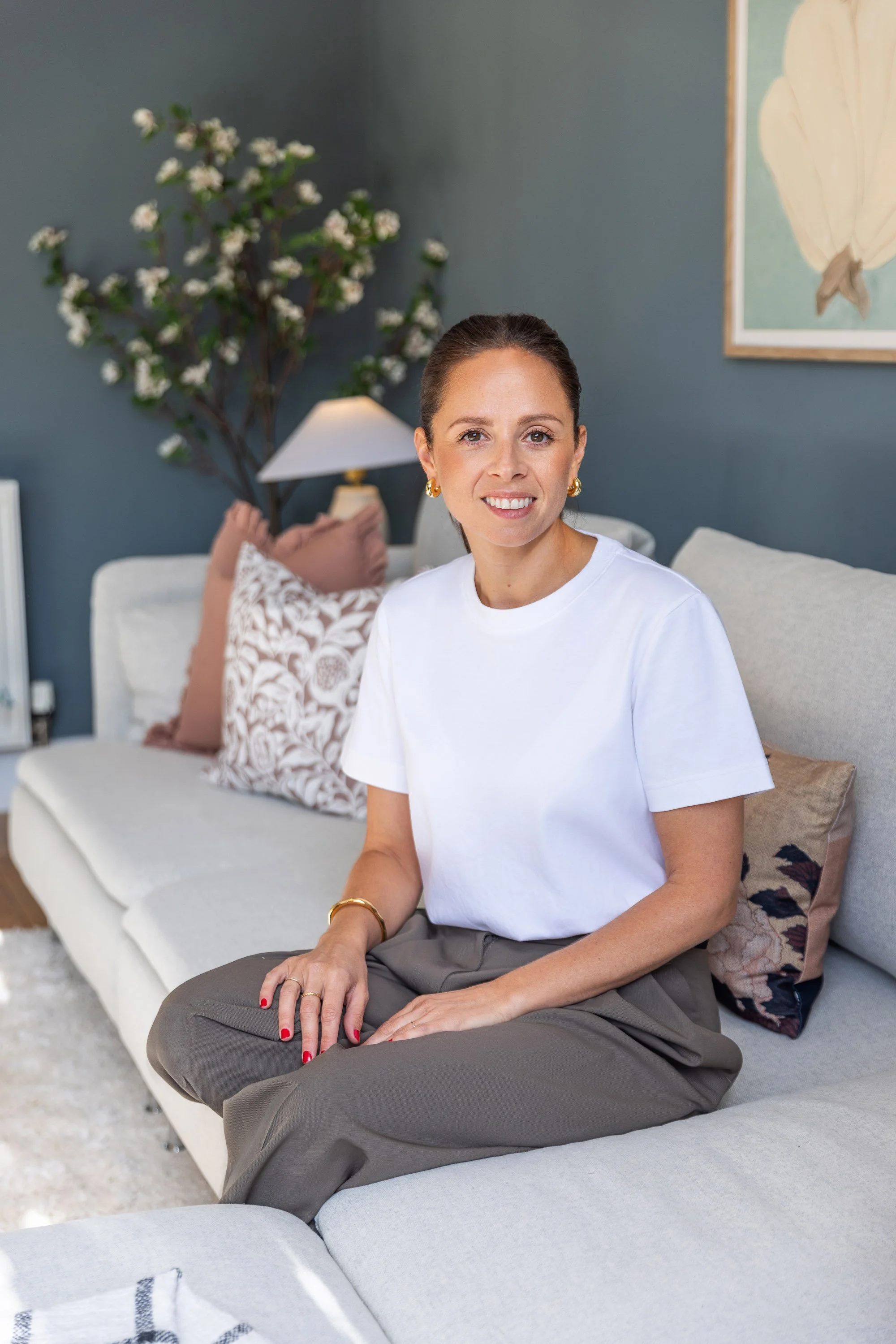 Woman sitting on a sofa with a relaxed posture, wearing a white t-shirt and olive green pants, with her hands resting on her lap, in a cozy living room with decorative pillows, a lamp, a wall art of a leaf, and a plant in the background.
