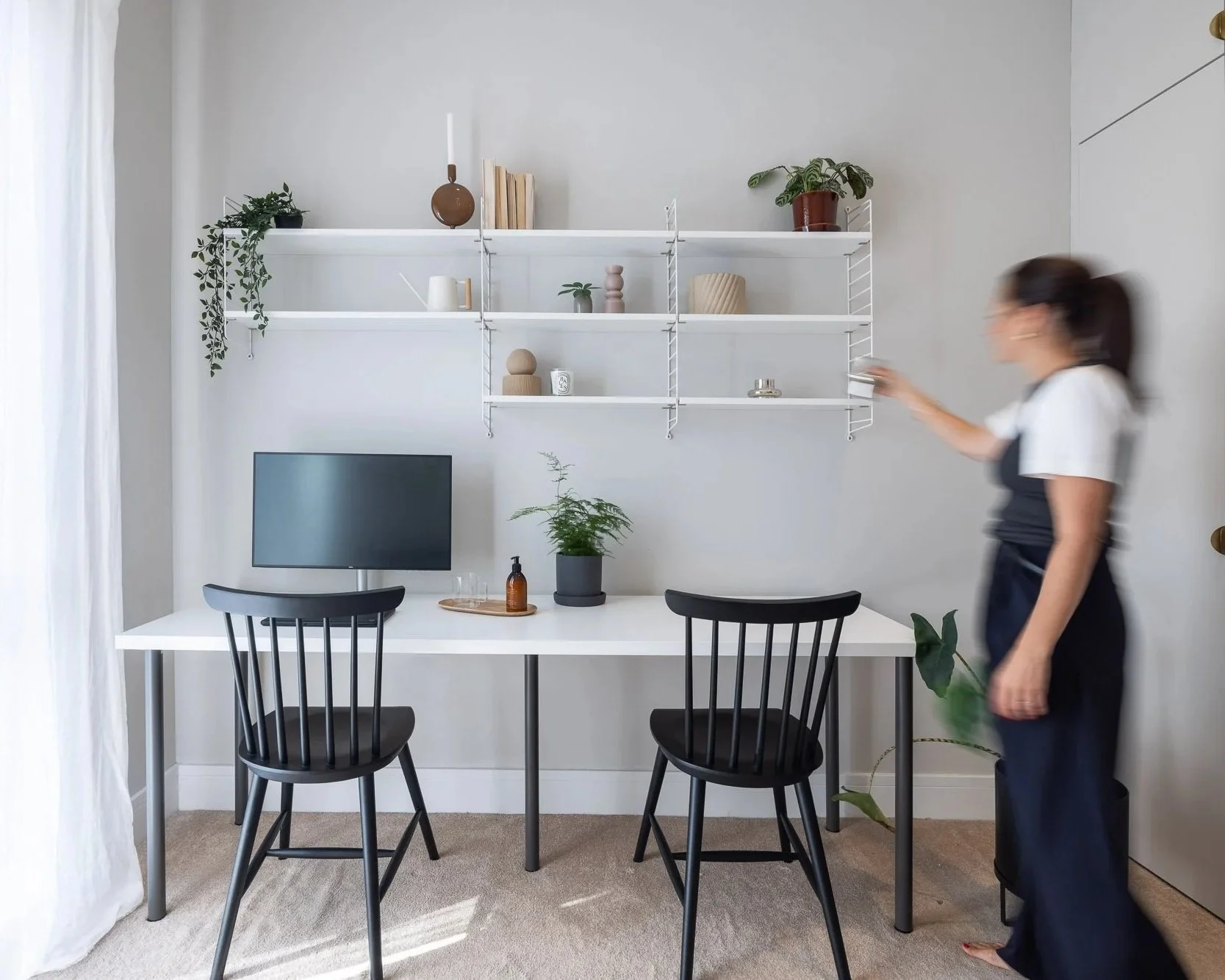 A woman standing in a minimalistic home office, next to a white desk with two black chairs, a monitor, and a plant. Behind her, wall-mounted white shelves hold decorative items including books, vases, and potted plants.