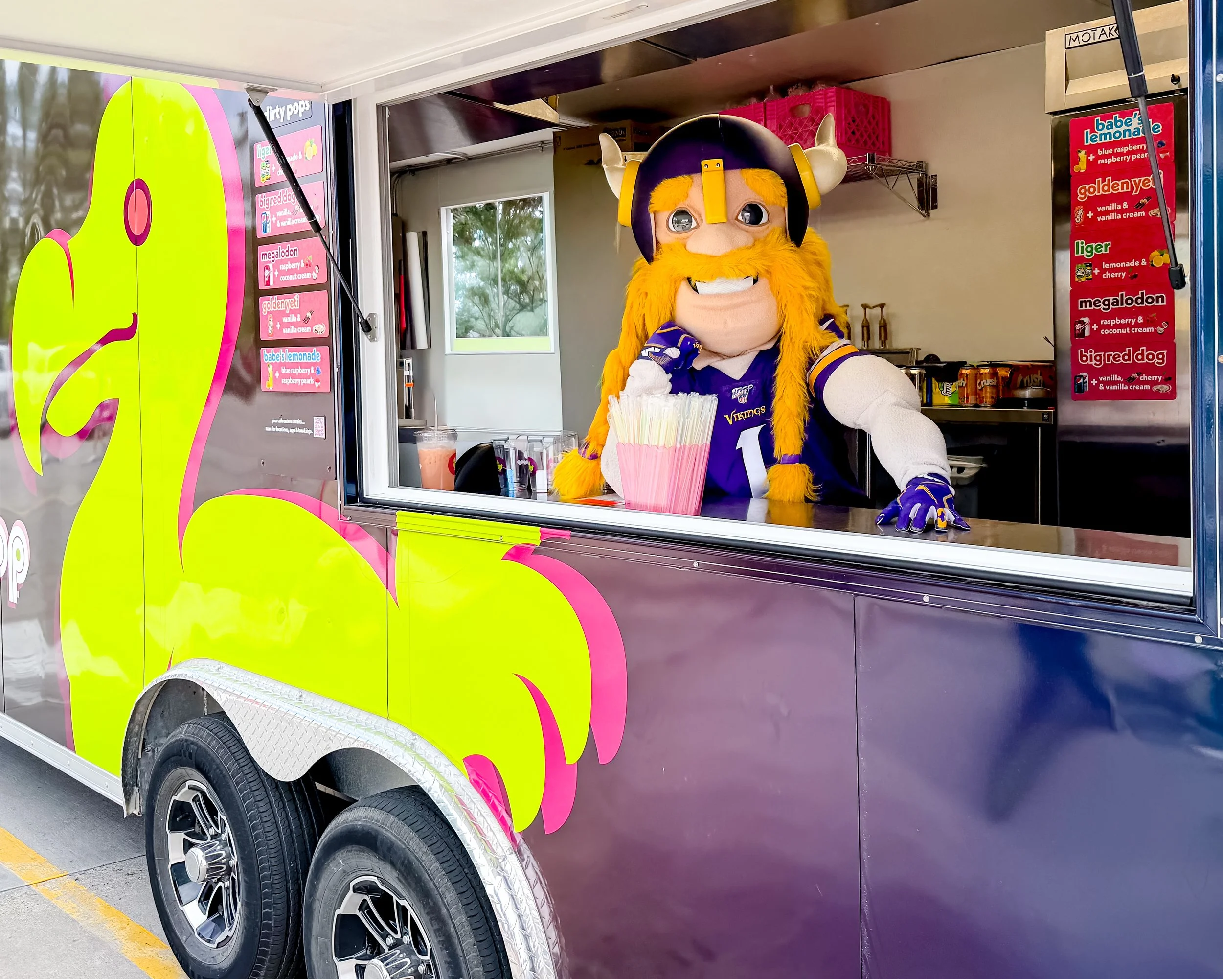 a person dressed as a viking mascot with a large helmet and orange beard, standing inside a colorful ice cream truck. the truck has pink, yellow, and purple decorations and a menu with drink flavors displayed on the side.