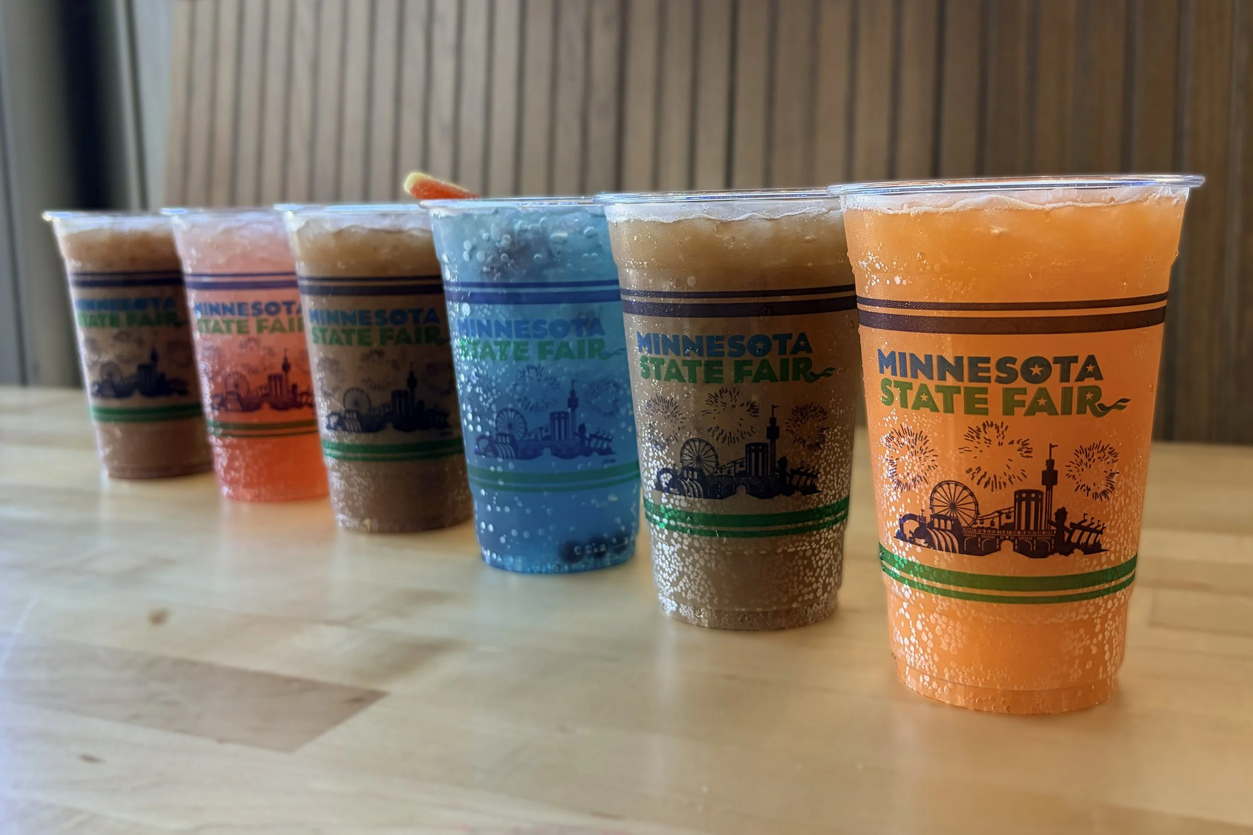 five plastic cups with colorful drinks labeled 'minnesota state fair' are lined up on a wooden surface, with a wood-paneled wall in the background.