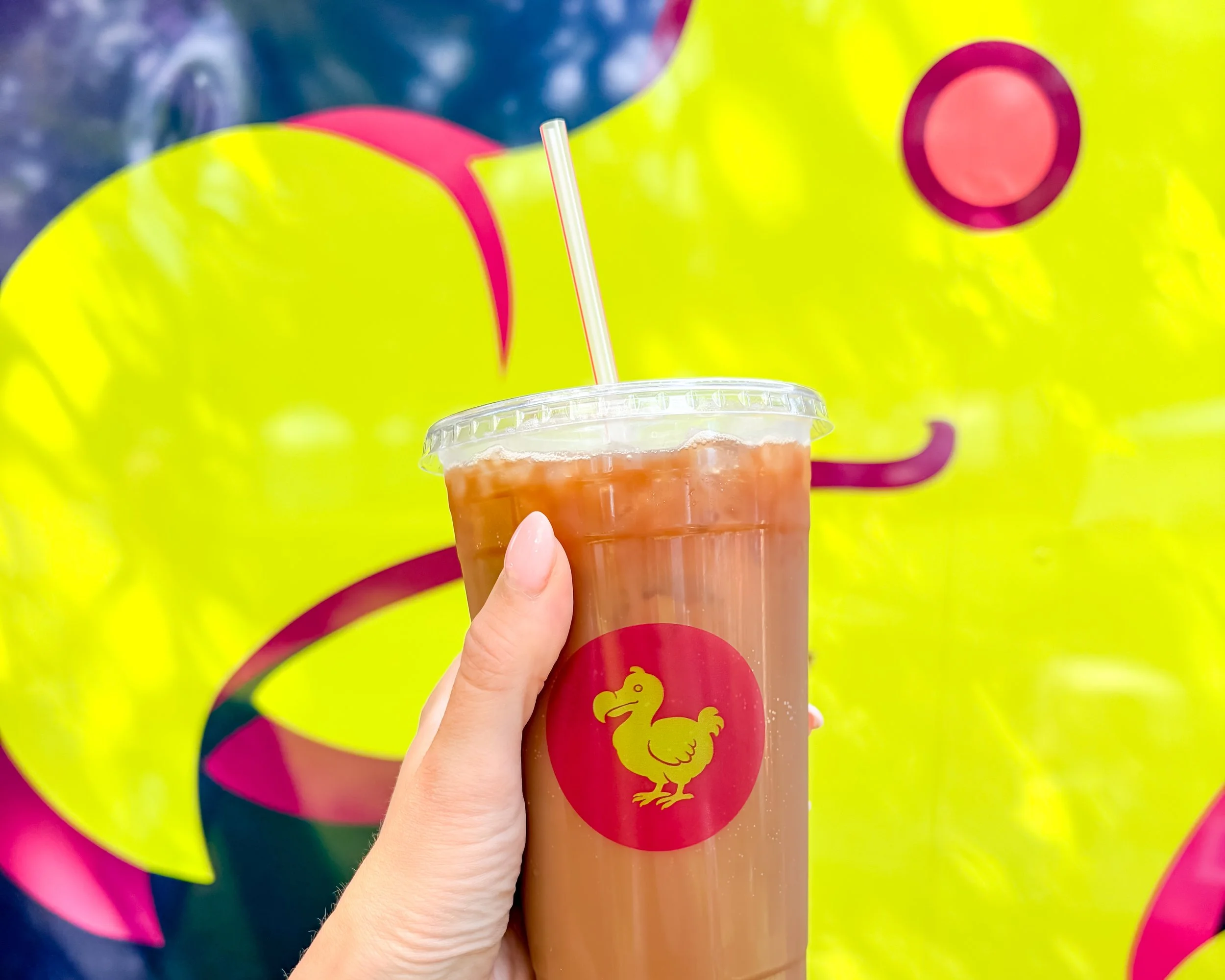 close-up of a hand holding a plastic cup with a brown beverage and a straw, with a colorful background featuring a lime green dodo bird.