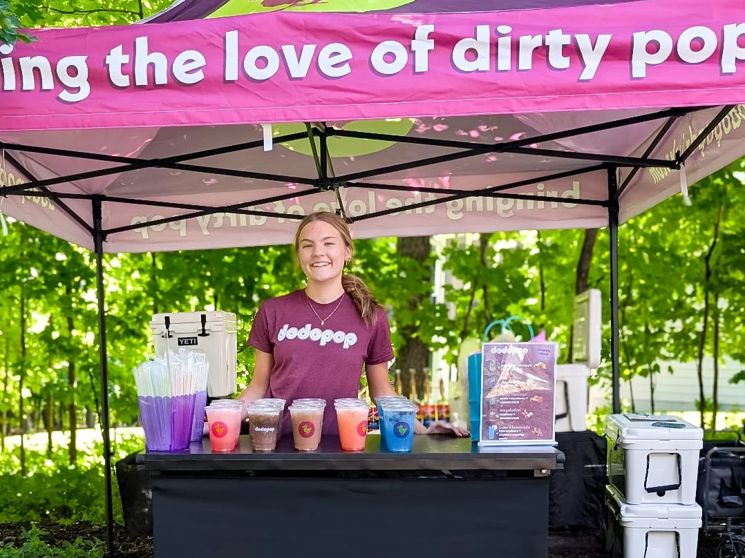 a young woman standing behind a colorful display of drinks at a pop-up stand under a pink canopy with the phrase "bringing the love of dirty pop". The stand has cups with various colorful beverages.