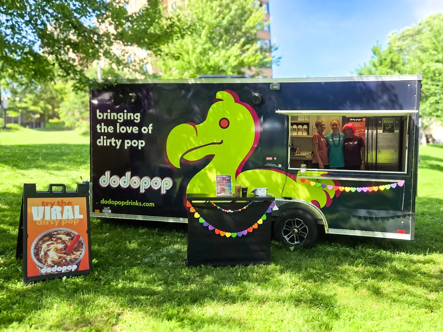 food truck with dark exterior and colorful branding, featuring a large lime green dodo bird and text promoting dirty pop. the truck is outside in a park with green grass and trees, and has a window with three people inside.